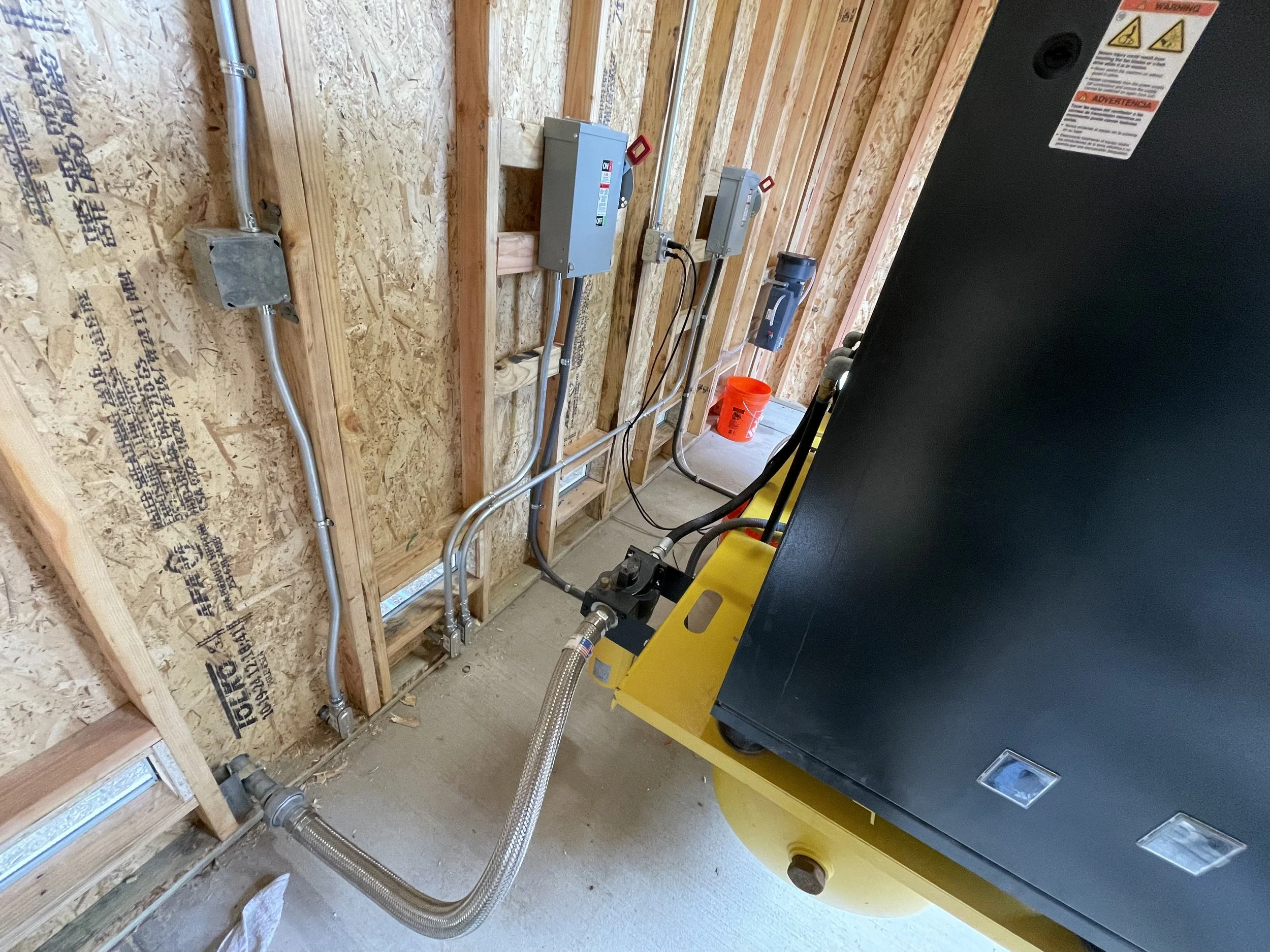 Construction room with exposed plywood walls, electrical wiring, and a piece of equipment, possibly a generator, with a yellow base.