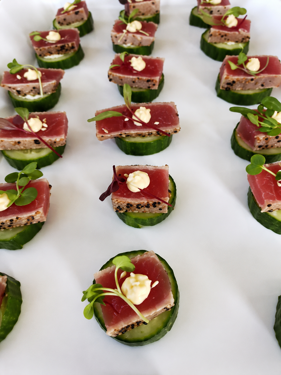 Cucumber slices topped with slices of raw fish, microgreens, lemon dollops, and creamy sauce, arranged on a white surface.