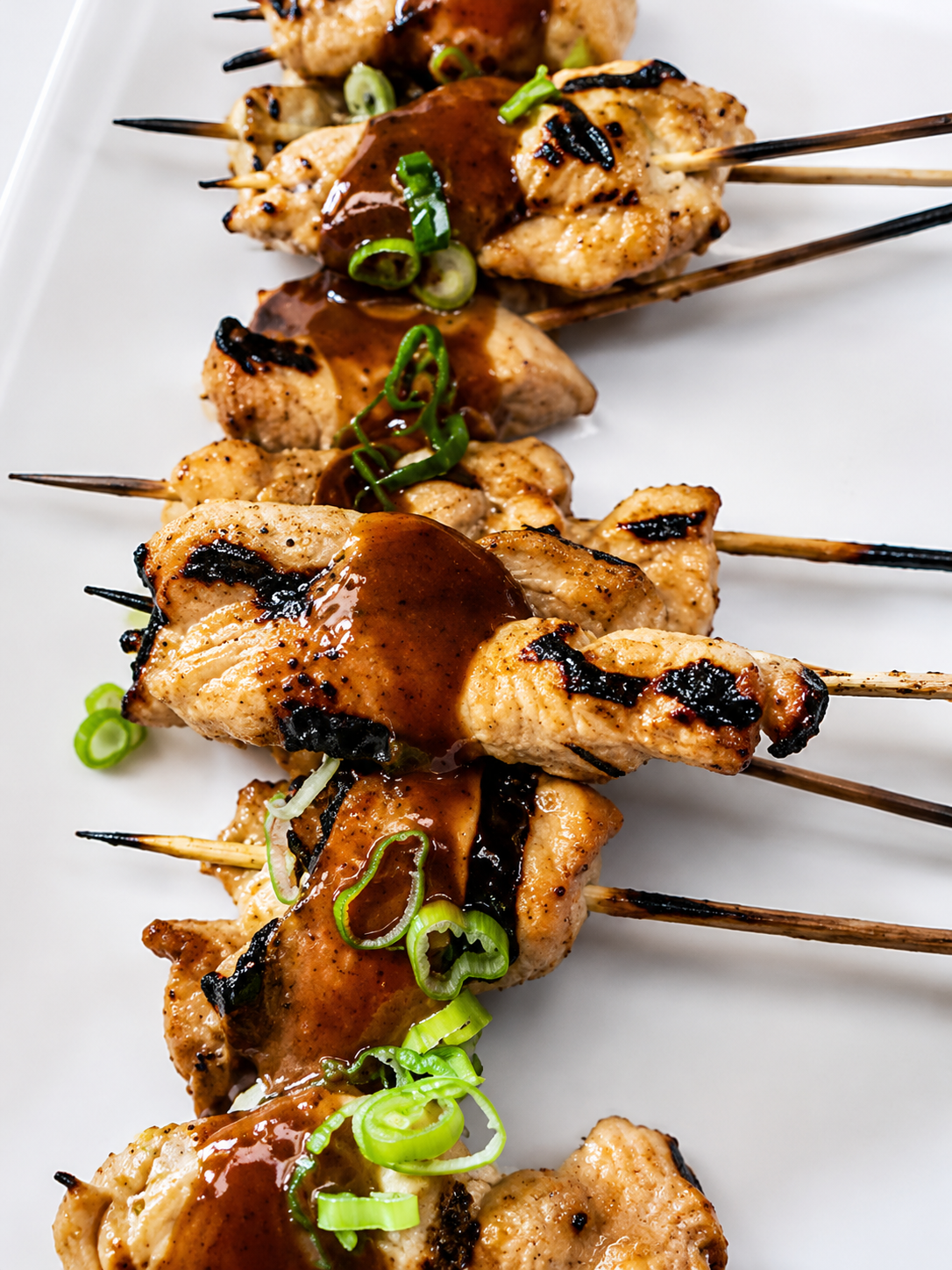 Close-up of grilled chicken skewers topped with green onions and dark sauce on a white plate.