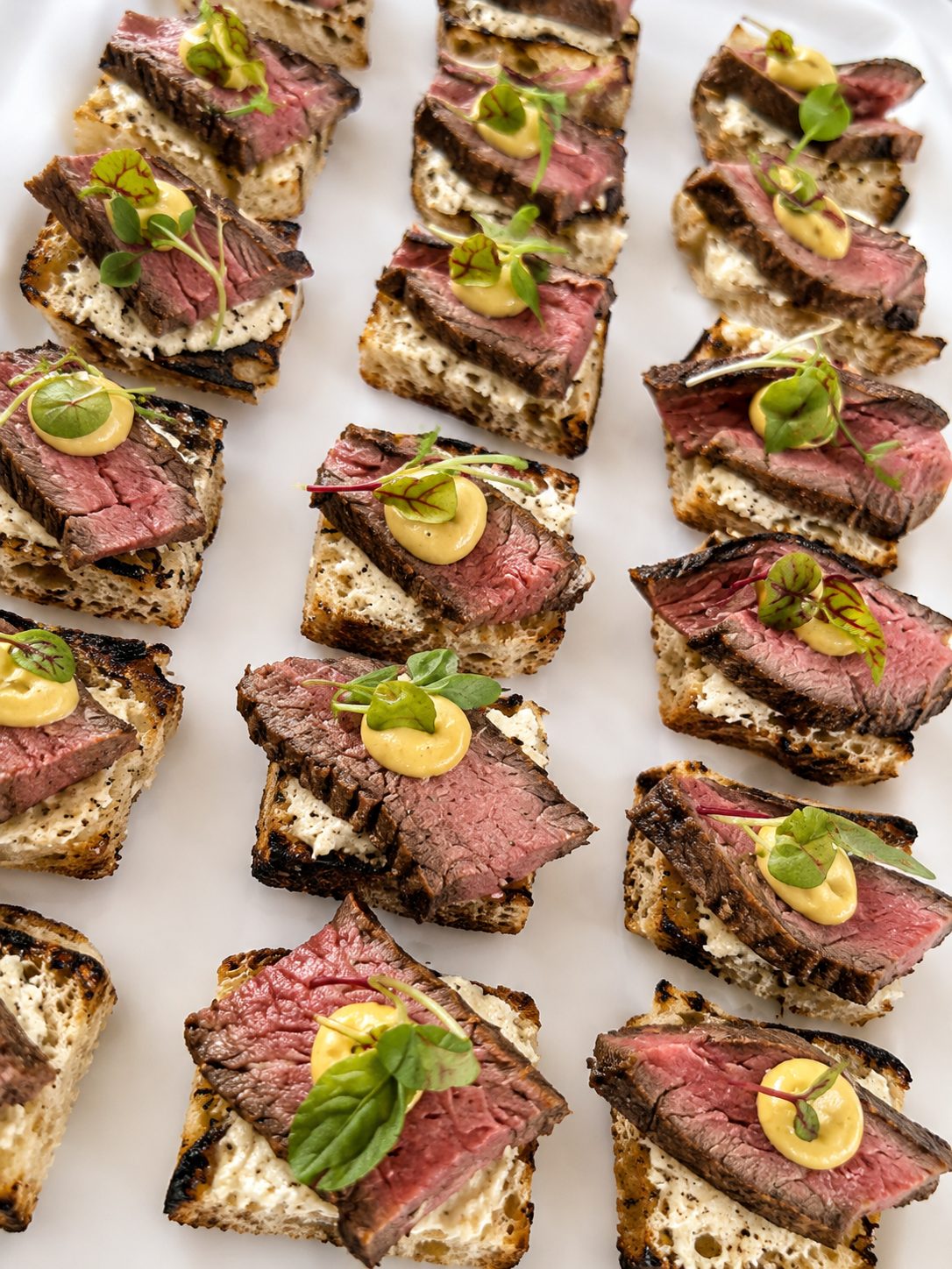 Slices of cooked steak on toasted bread with a dollop of yellow sauce and garnished with microgreens, arranged on a white platter.