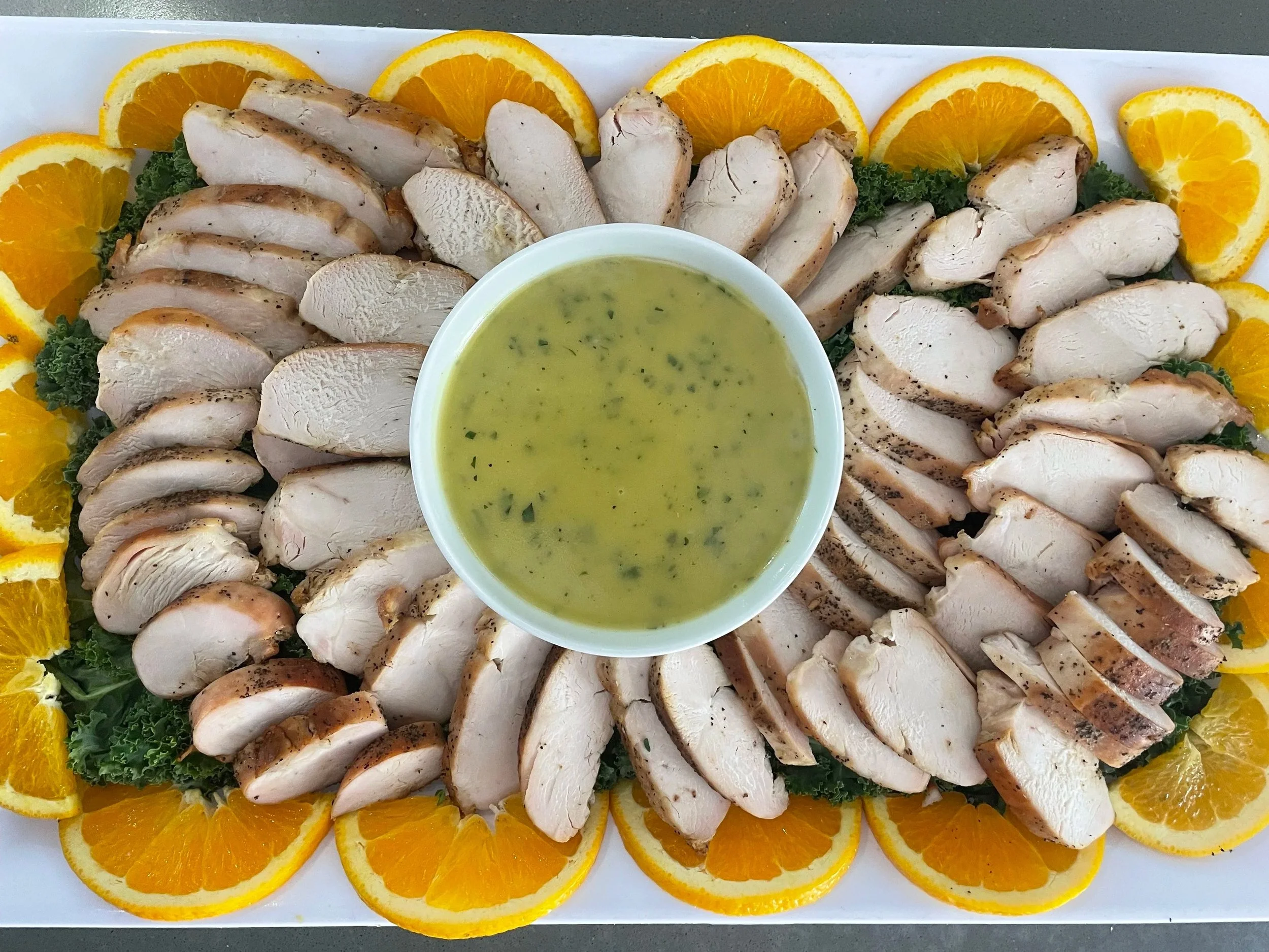 Sliced cooked chicken breast arranged on a platter, garnished with orange slices and leafy greens, with a small bowl of green herb sauce in the center.