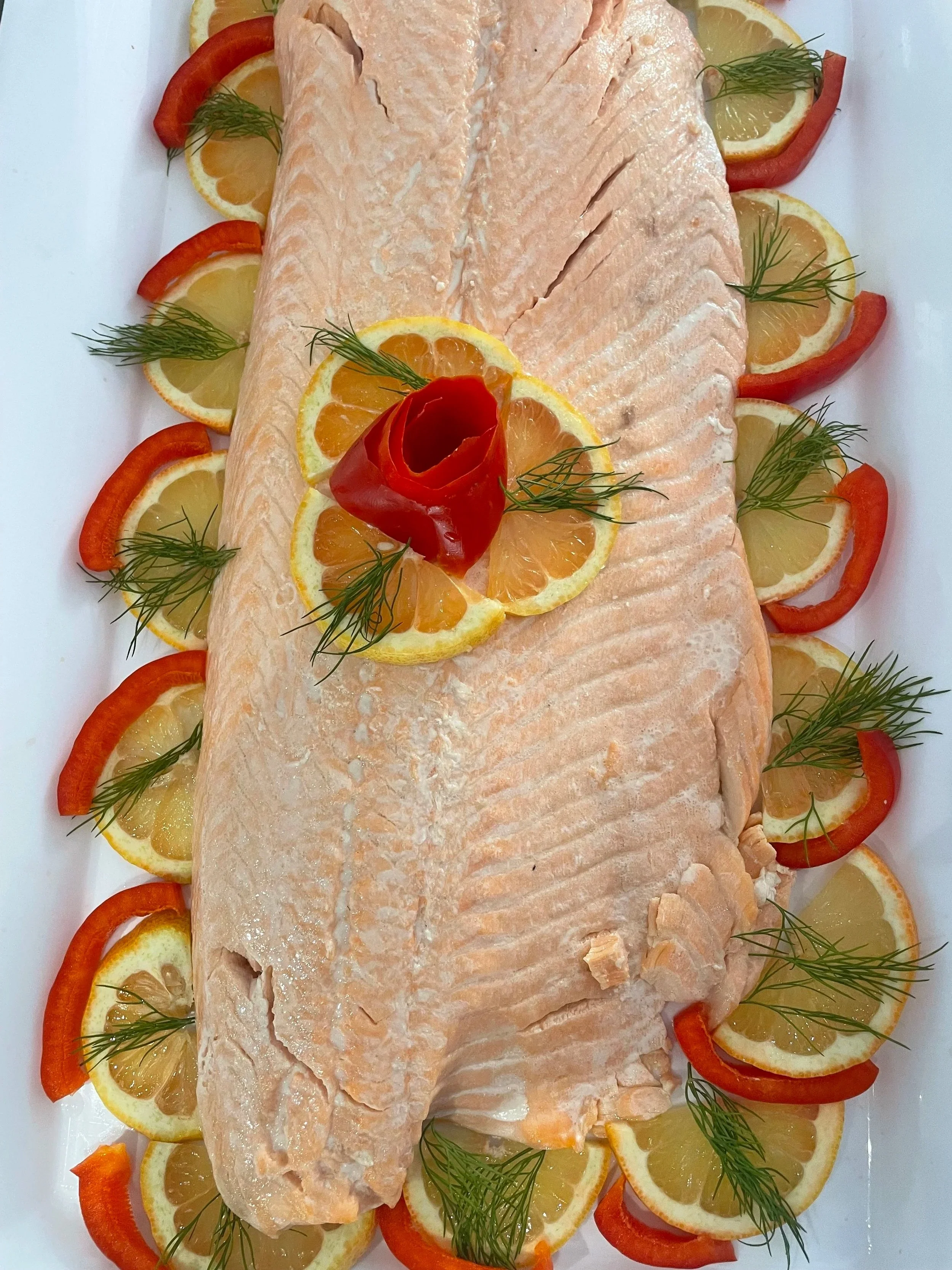 A cooked whole salmon fish garnished with lemon slices, red bell pepper pieces, and sprigs of dill, arranged on a white platter.