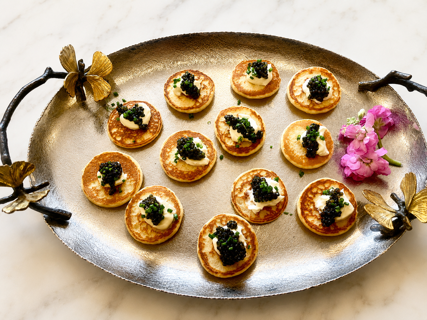 Small breakfast appetizers on a decorative tray with golden butterfly handles, garnished with cream, caviar, and chopped green onions, accompanied by pink flowers.