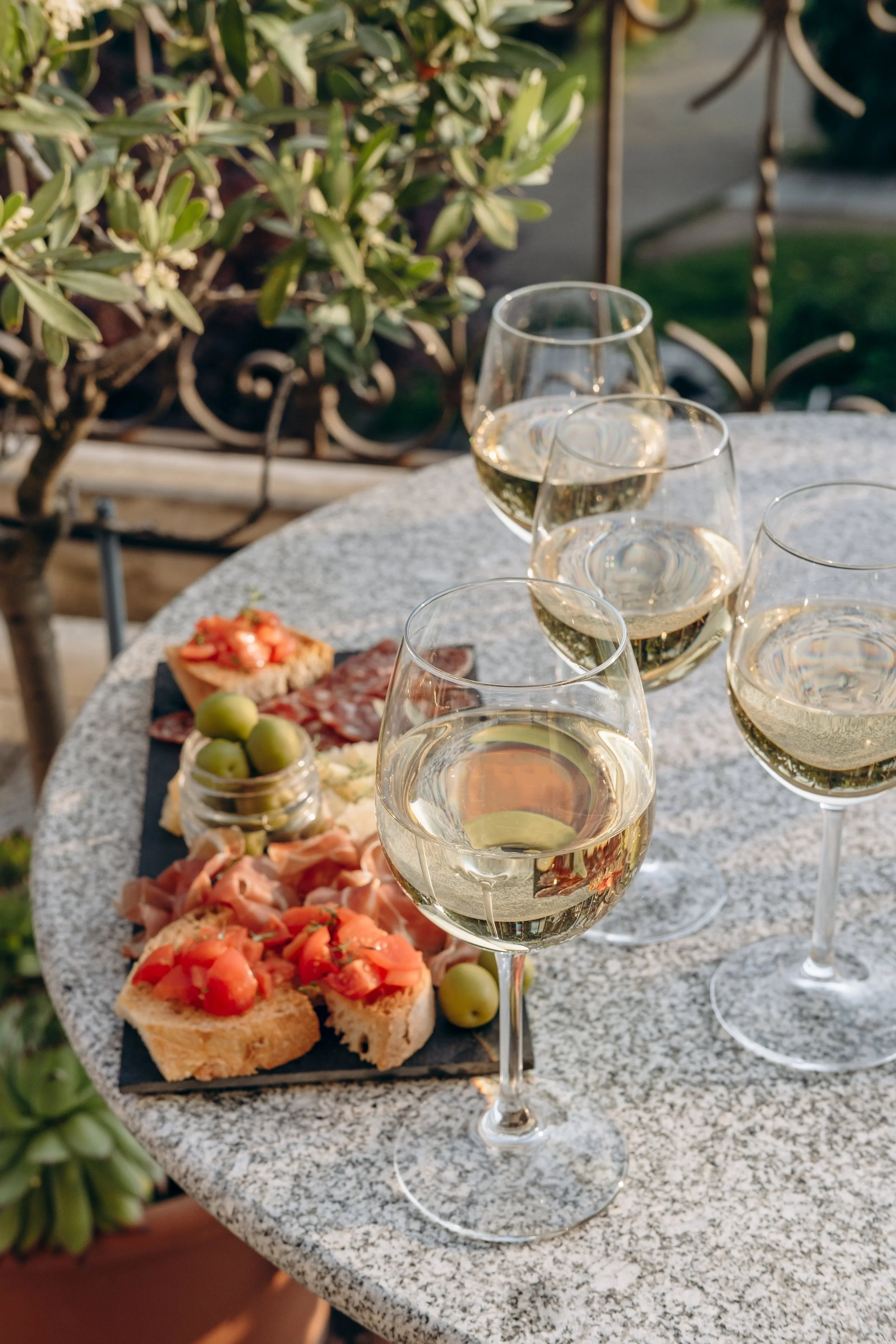 Four glasses of white wine on a round outdoor table with an assorted platter of appetizers, including sliced bread with chopped tomatoes, cured meats, green olives, and cheese. Green plants and garden decorations are visible in the background.