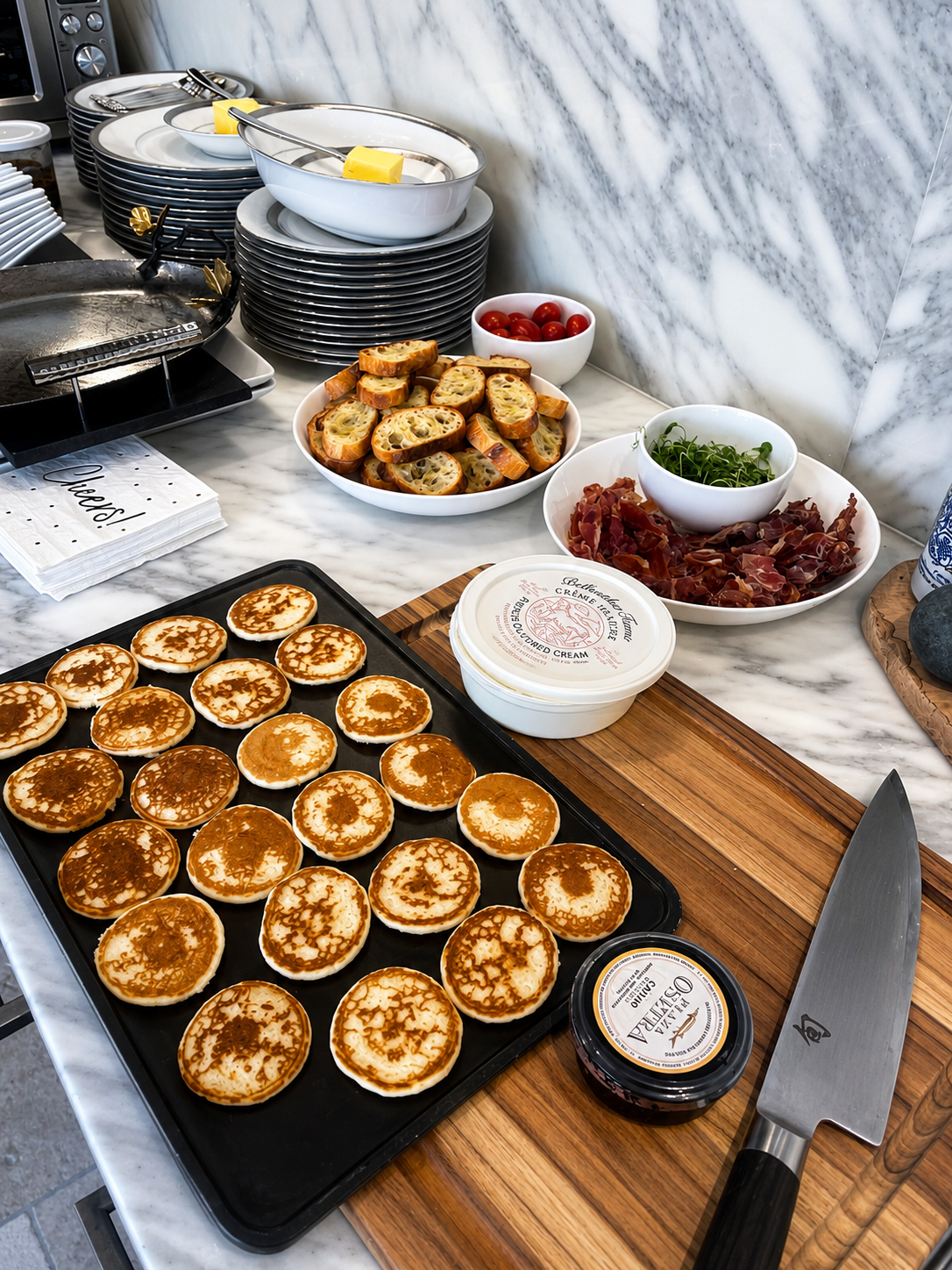 Tray of small pancakes or blinis on a black baking sheet, with various toppings and accompaniments on a marble countertop, including bread slices, cherry tomatoes, sliced cured meats, greens, butter, cream, and a knife.