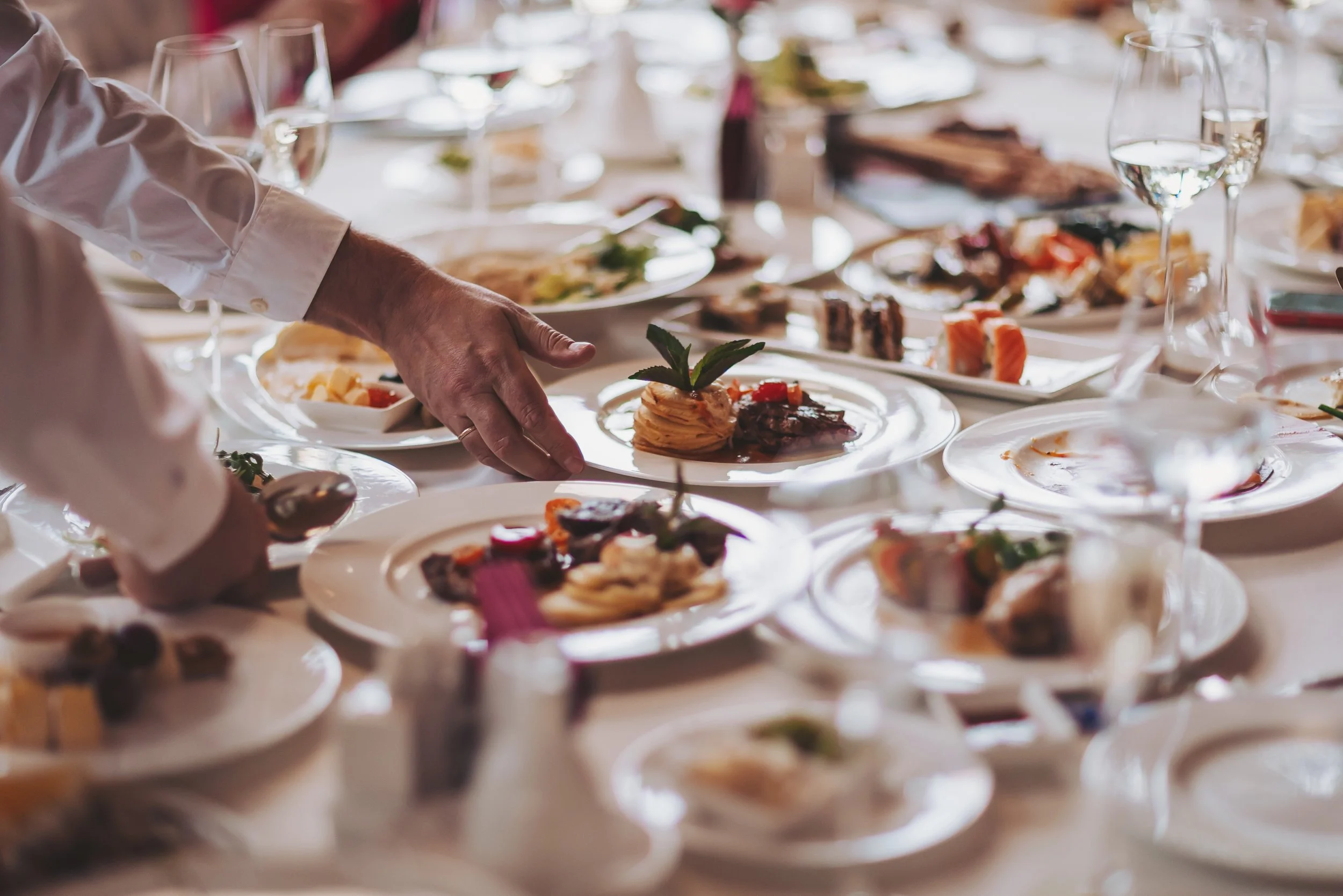 A close-up of a person's hand reaching for a plate of assorted food on a table filled with various dishes, glasses, and tableware at a social gathering or event.