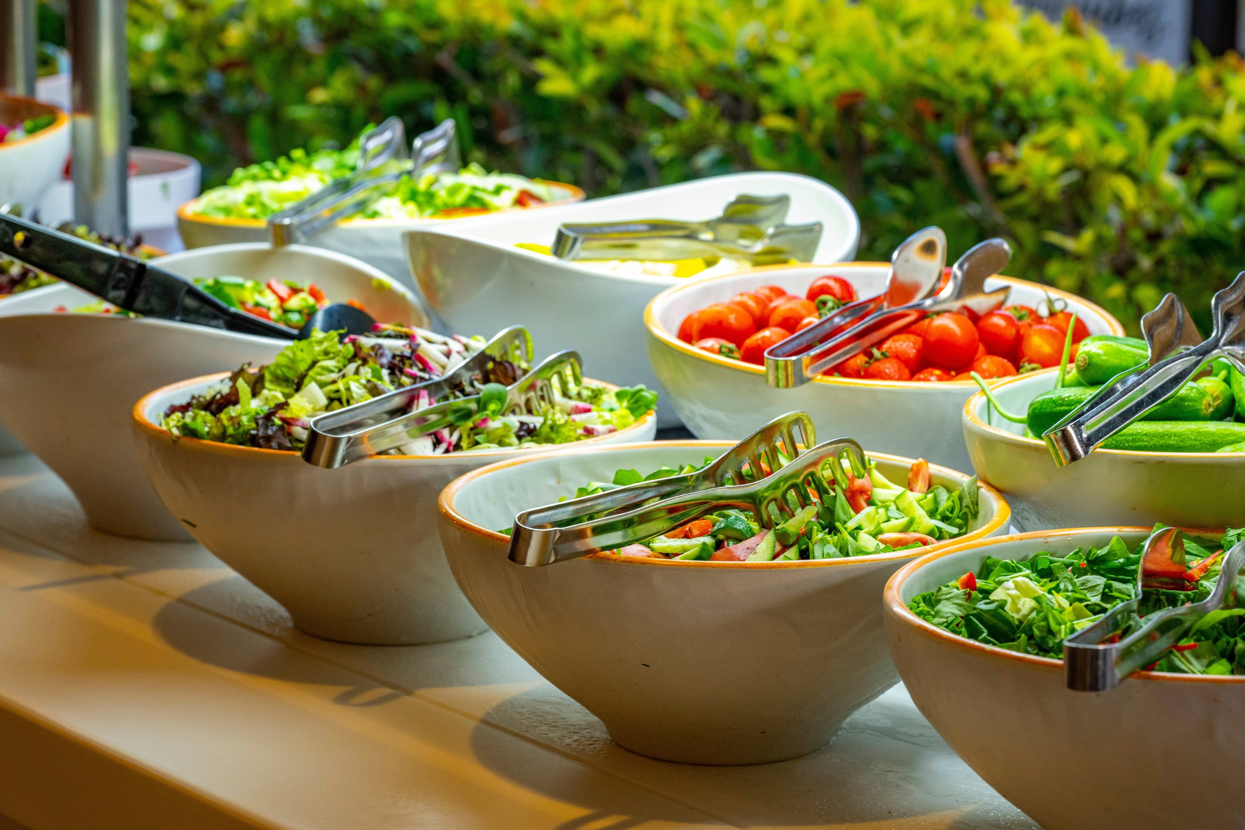 A buffet of fresh salads and vegetables in white bowls with tongs, set against a background of green bushes and outdoor setting.