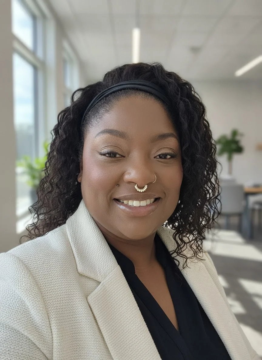 A woman with curly hair wearing a black headband, a nose ring, and a white blazer, smiling in a well-lit office with large windows and plants in the background.