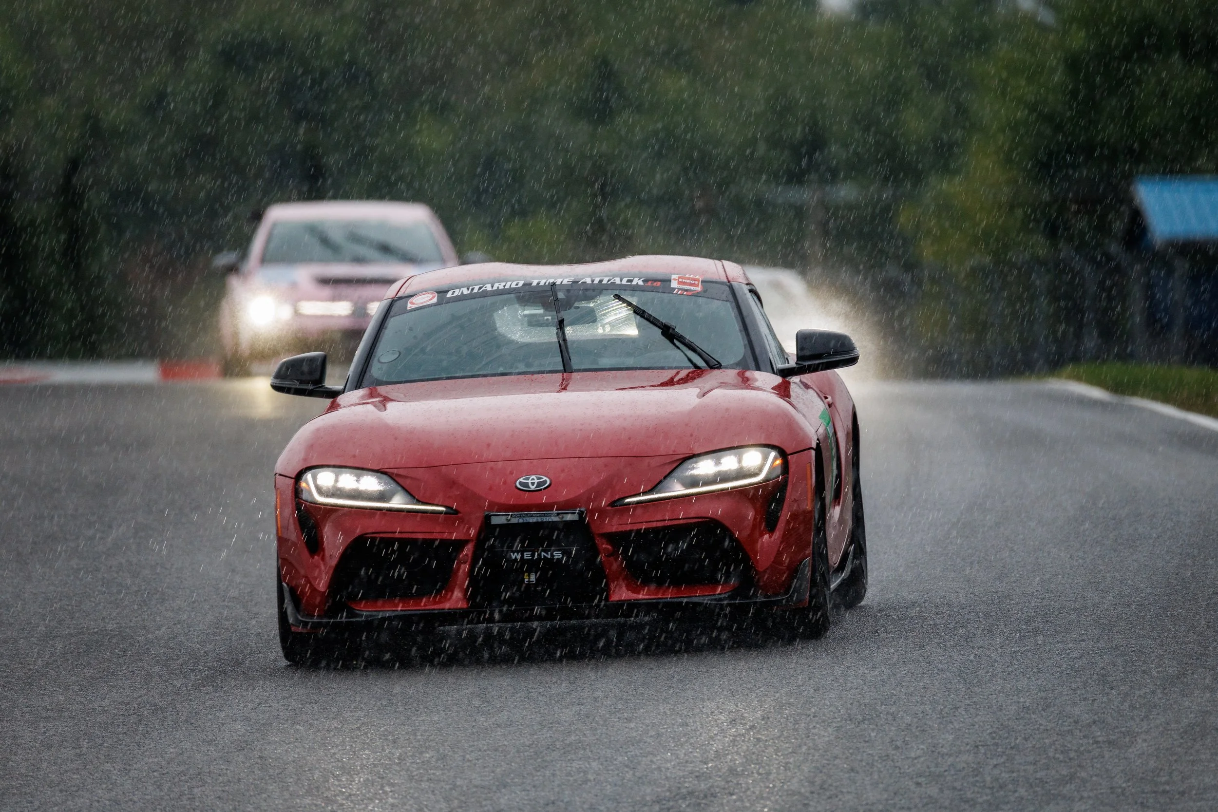 Red sports car driving on wet road in the rain, with a white vehicle behind it and a blue structure to the right.