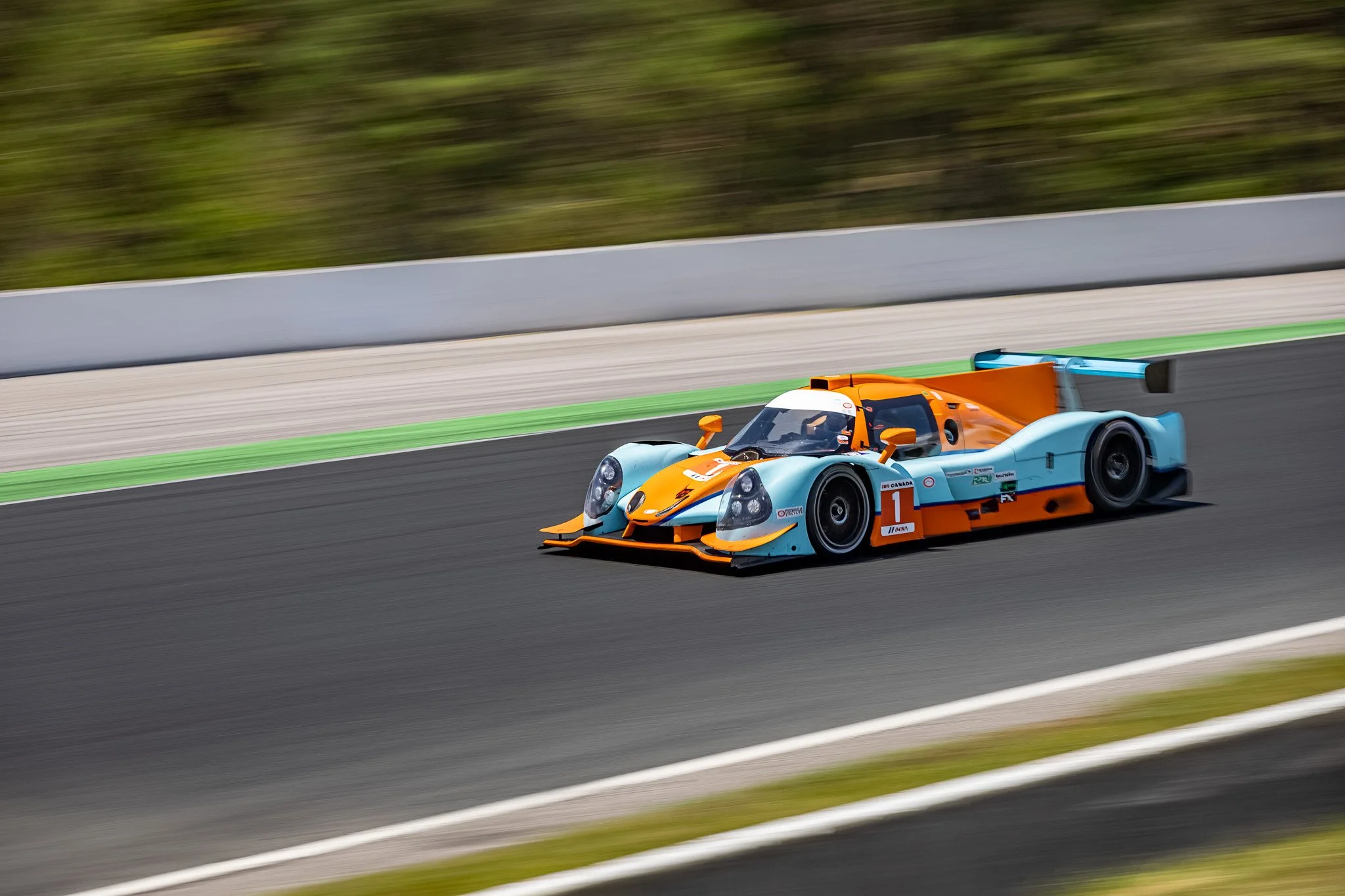 A race car in orange and blue speeding on a racetrack with a blurred background of greenery.