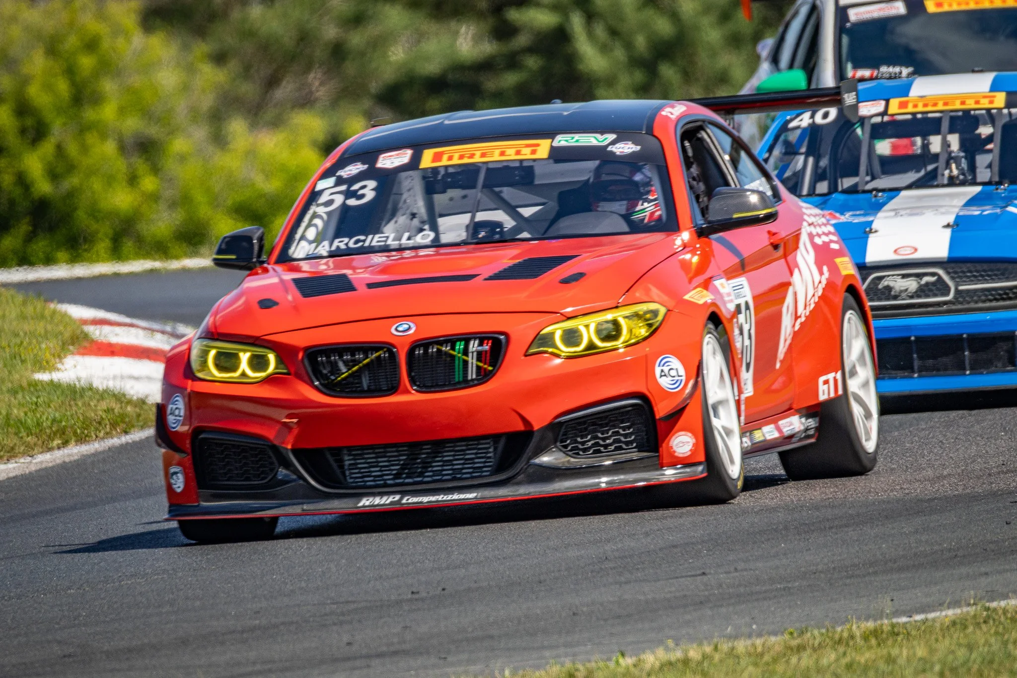 A red BMW race car with the number 53 and the name Marciello on the windshield is on a race track, with a blue and white car close behind. The cars are navigating a curve on the race track surrounded by greenery.