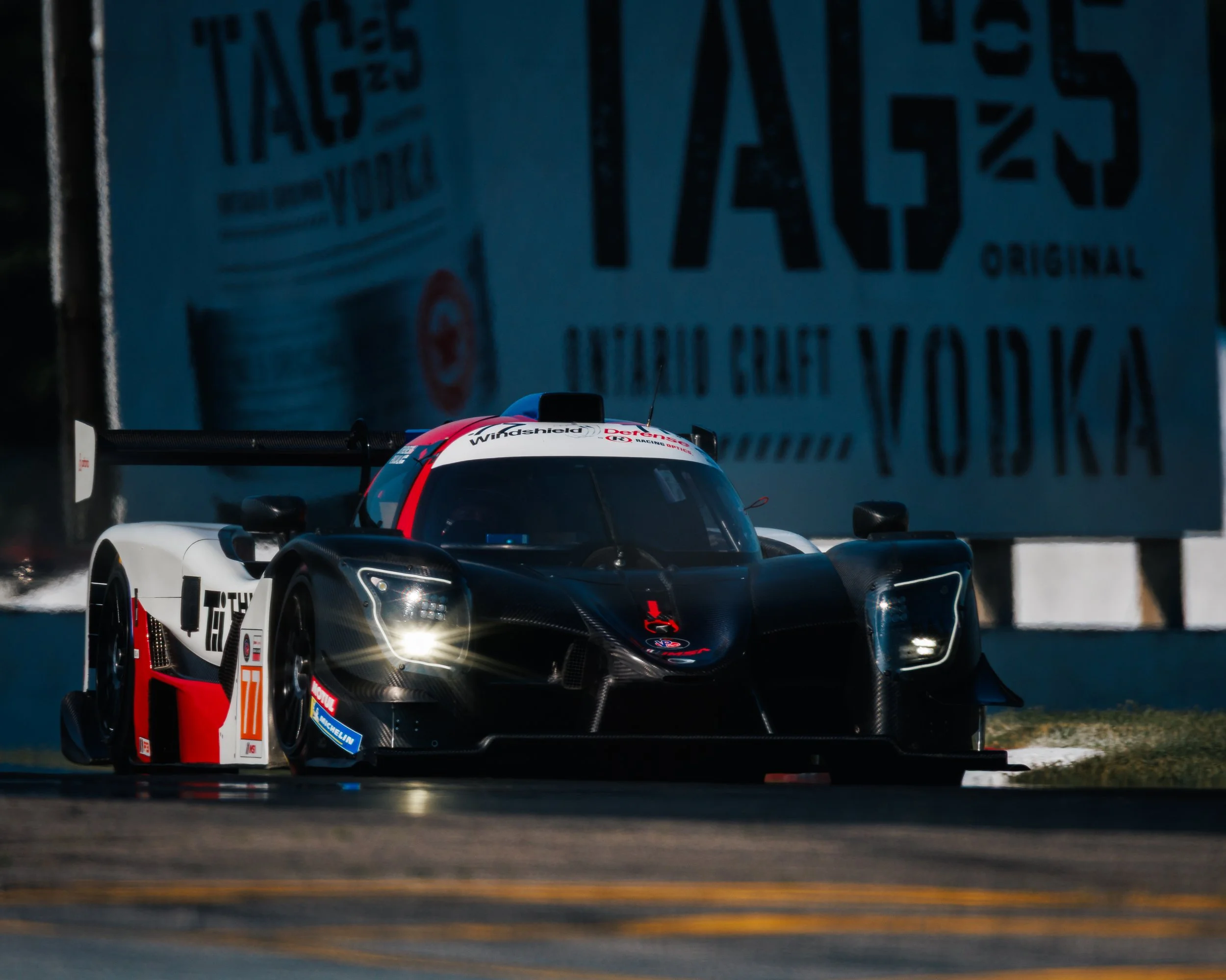 A race car on a track during a race. The car has sponsorship logos and a sleek, aerodynamic design.