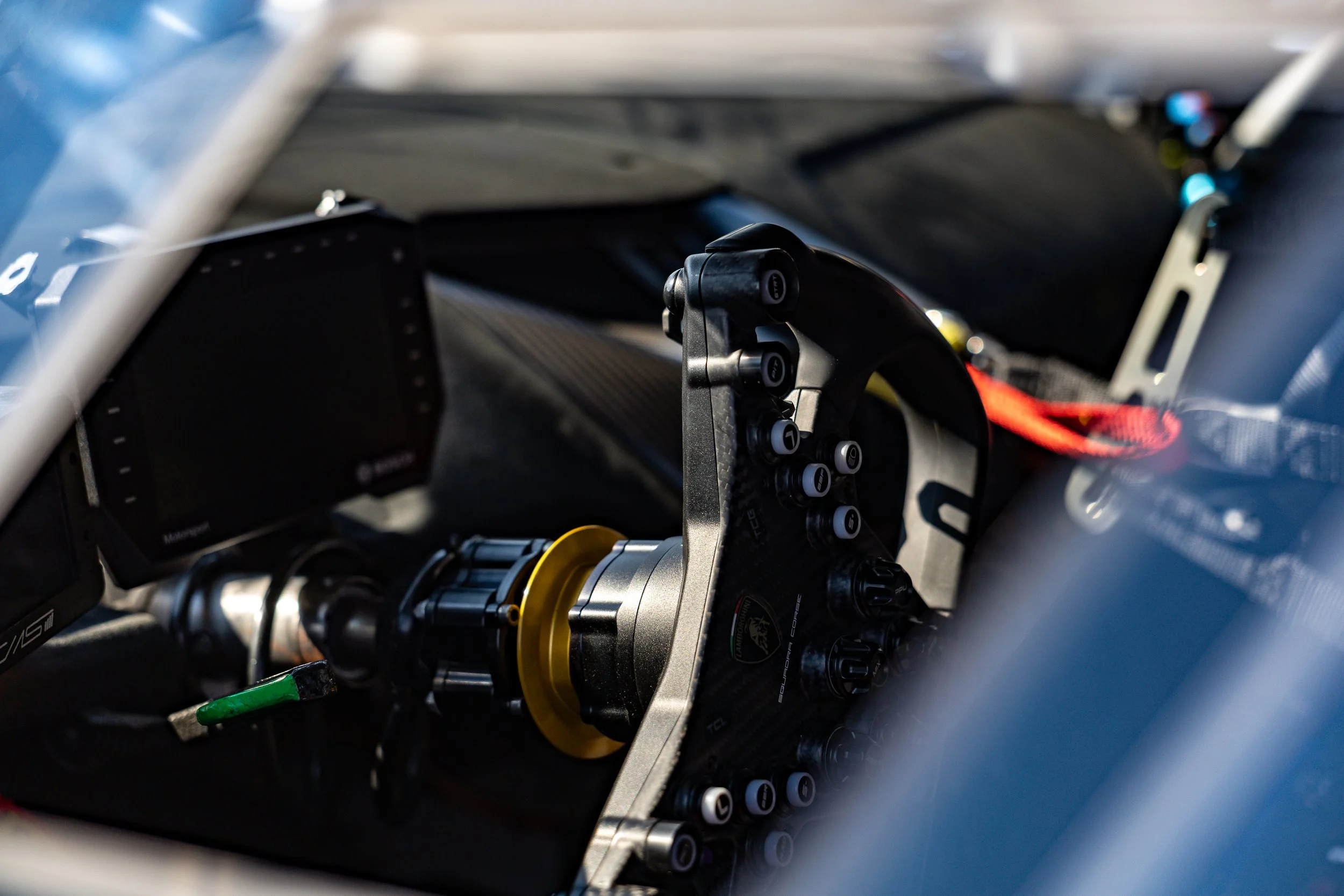 Close-up of a car racing car's interior, showing a steering wheel with multiple buttons and a label reading 'Derek,' and part of the dashboard and electronic equipment.