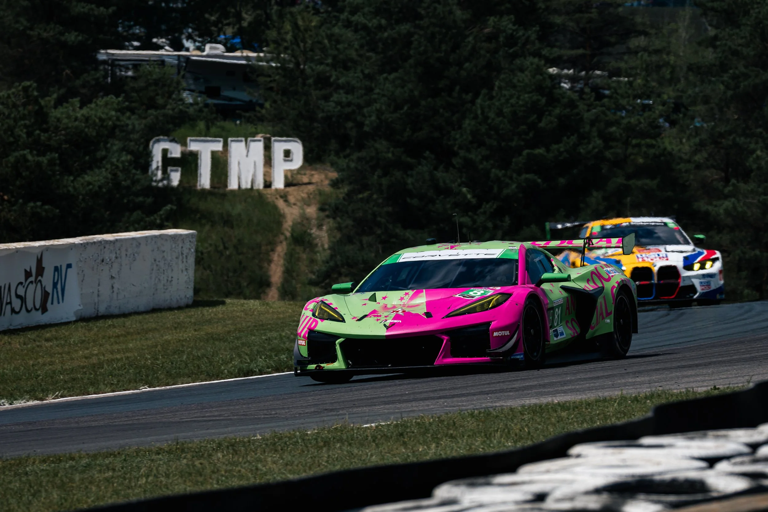 Race cars on a track, including a pink and green car in the foreground and a white car with colorful logos behind, with a sign displaying the letters 'CTMP' and trees in the background.