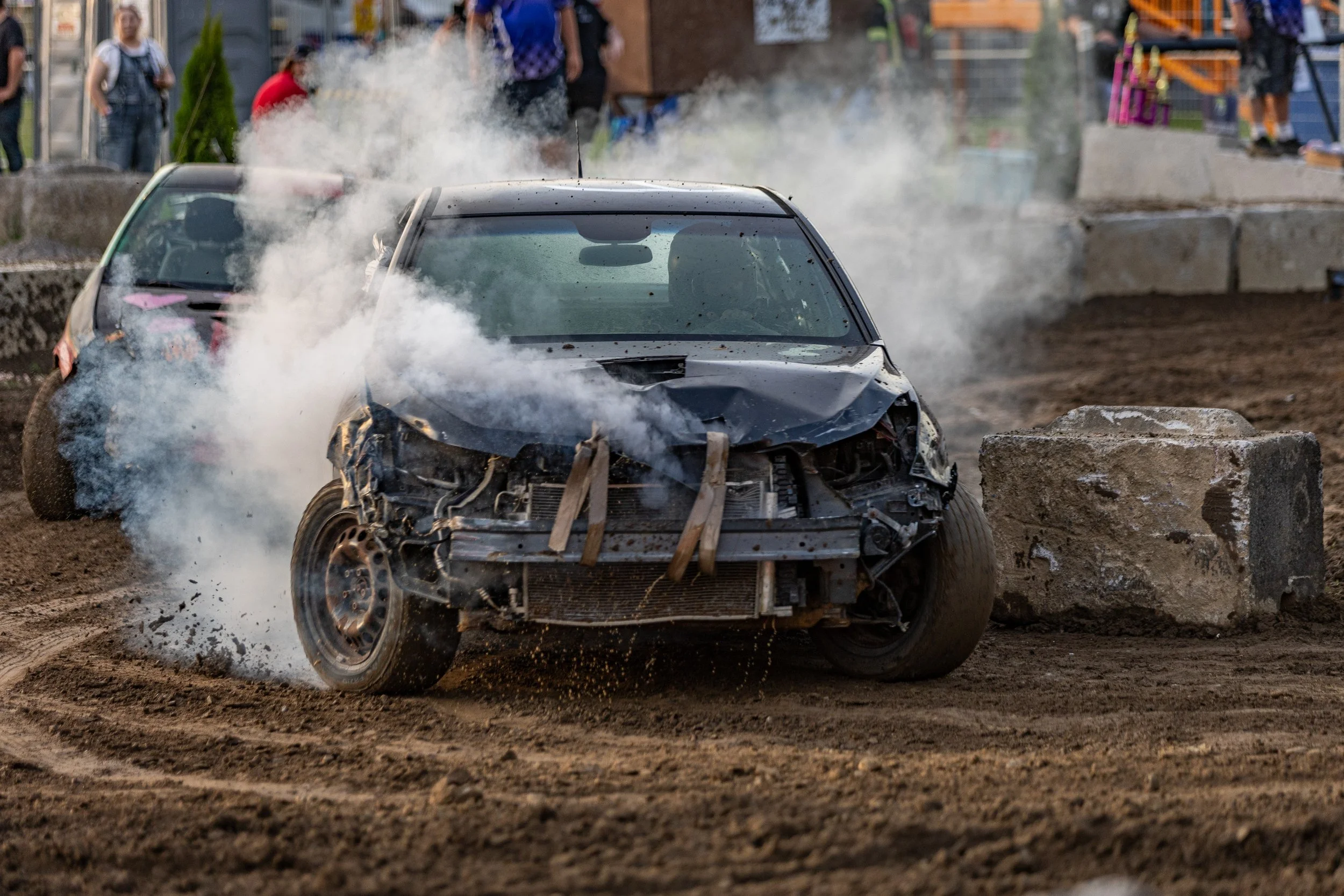 A damaged black racing car on a dirt track, with smoke and debris around it during a race event.