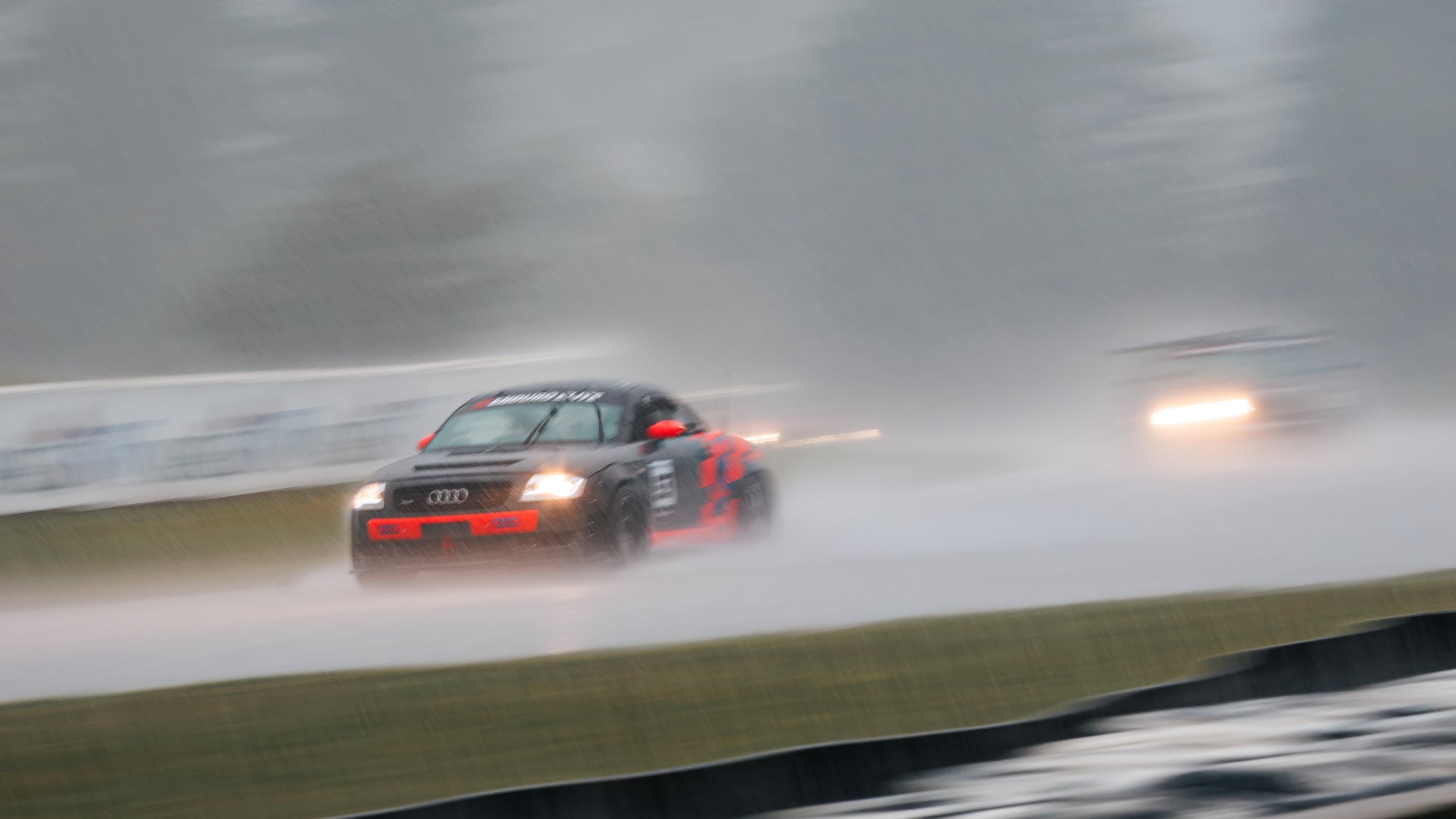 A black race car with red accents speeding on a wet racetrack during a rainstorm, creating a spray of water behind it.