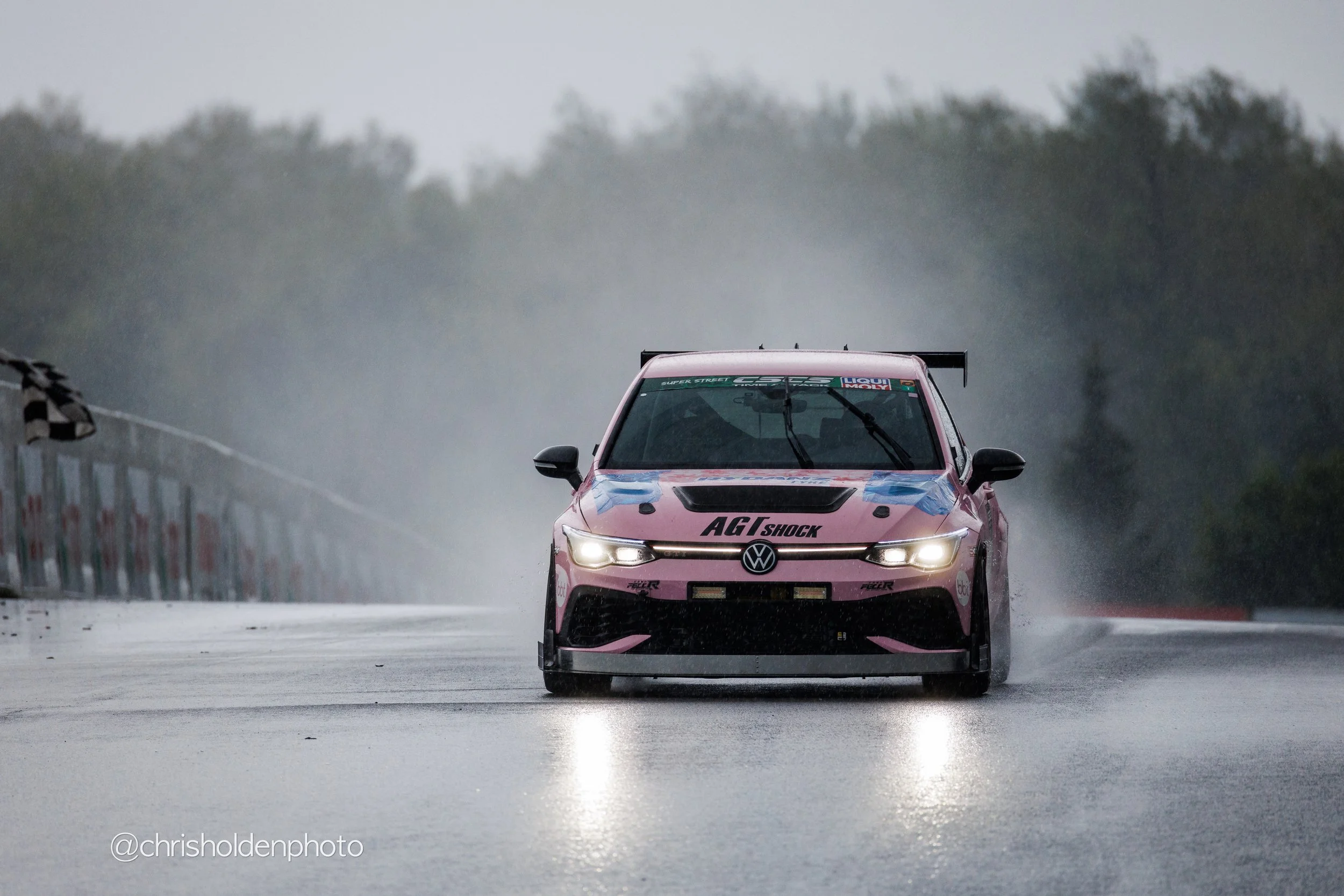 A pink race car with various sponsor logos, including Volkswagen and AGT Shock, driving on a wet race track through foggy weather, with trees in the background and a checkered flag on a pole to the left.