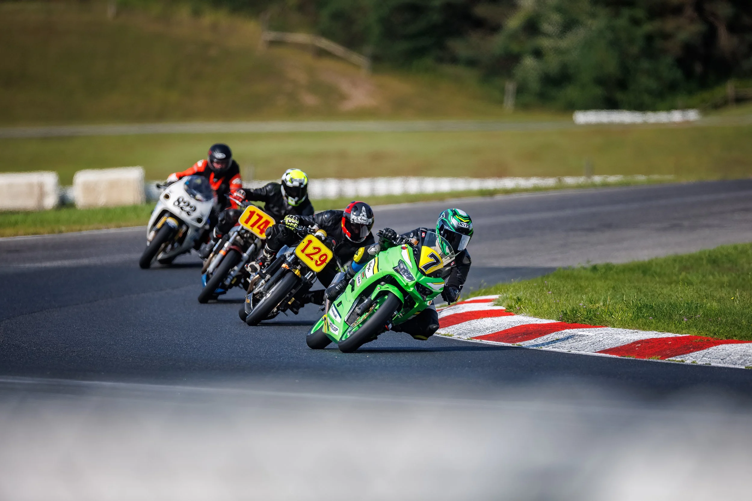 Motorcycle racers leaning into a turn on a race track, with the leading rider on a green motorcycle wearing a blue helmet, followed by riders on black motorcycles with yellow numbers and a white motorcycle with a yellow number.