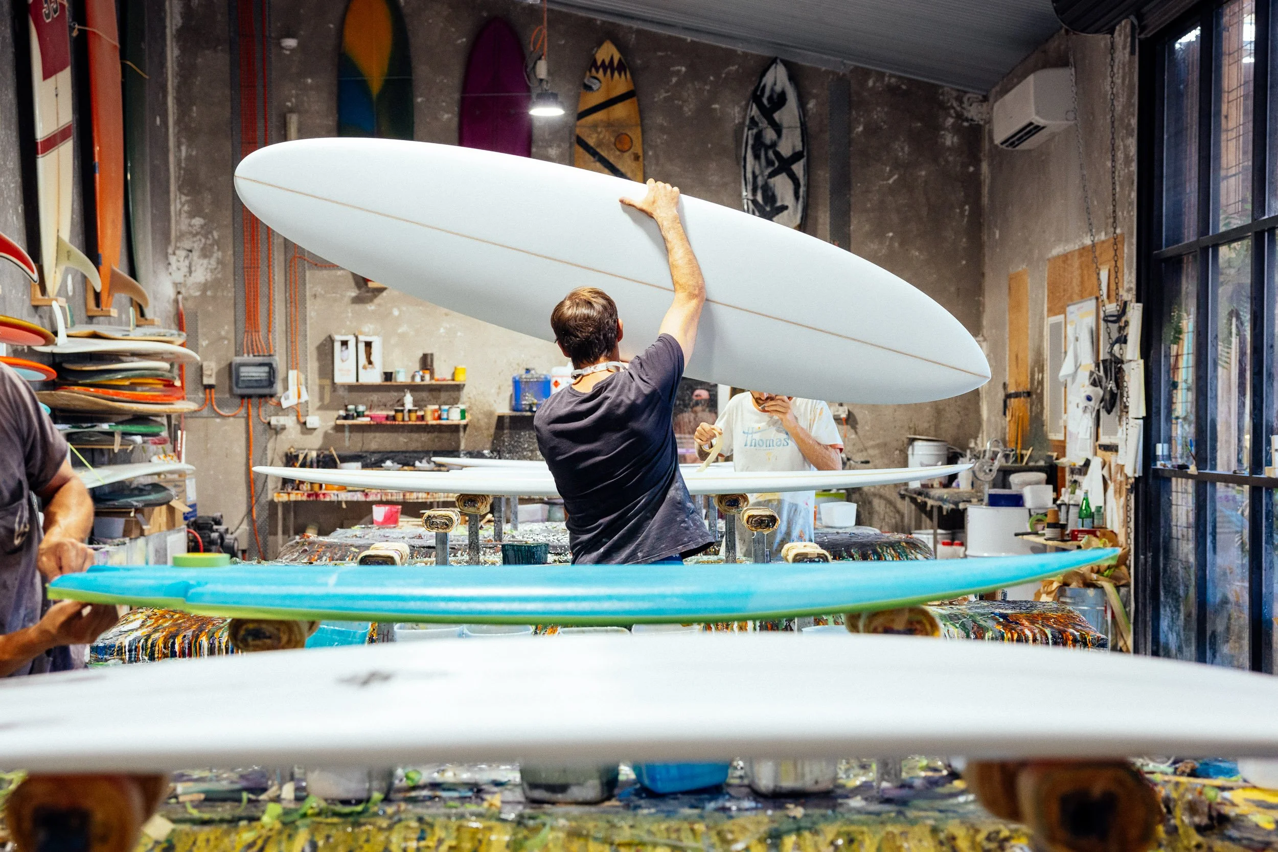 Surfboard workshop with people shaping and painting boards, various surfboards on display, tools and materials visible, creative workspace atmosphere.