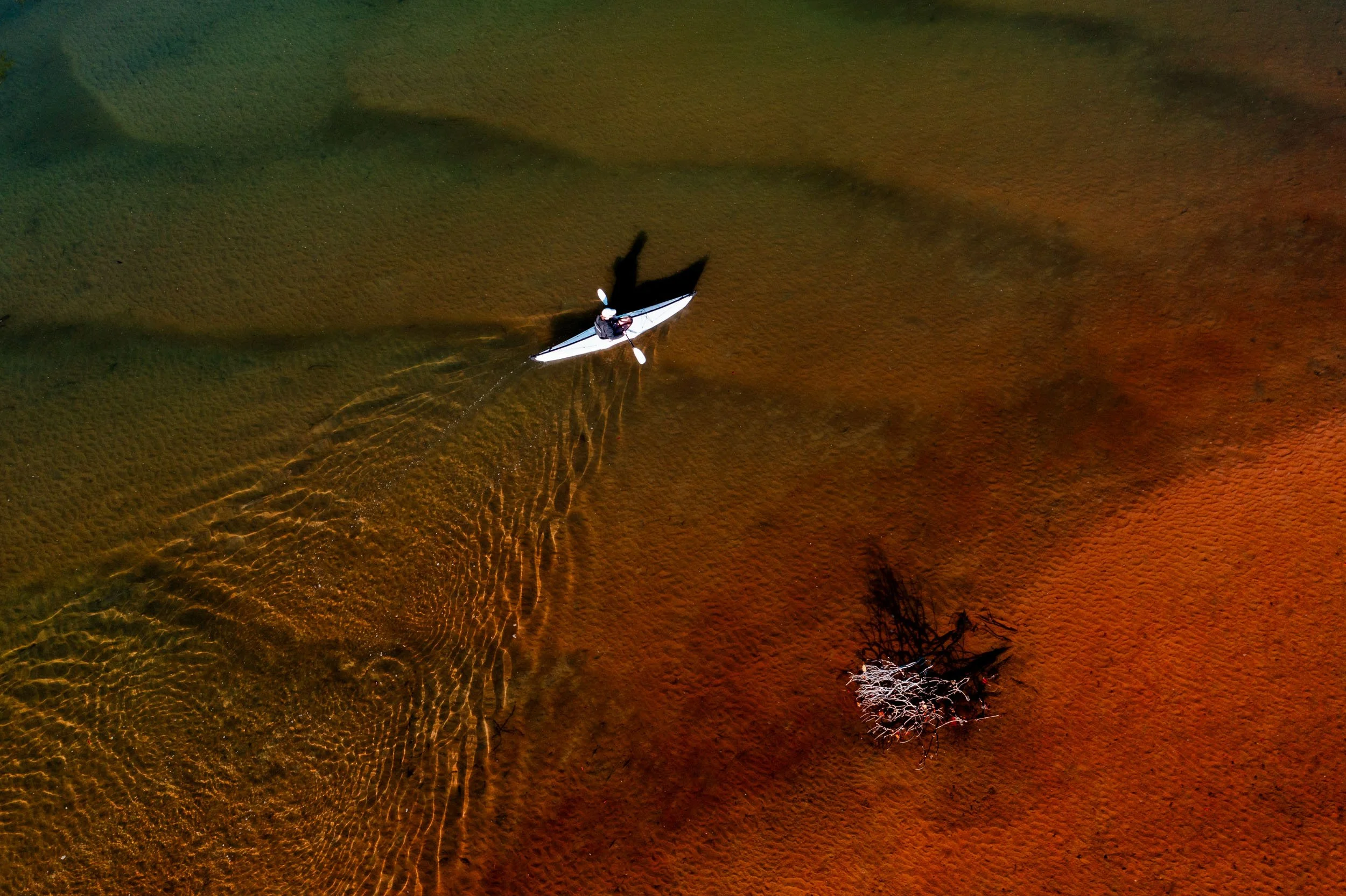 Aerial view of a person kayaking in shallow water with visible sand patterns, alongside a small cluster of submerged branches.