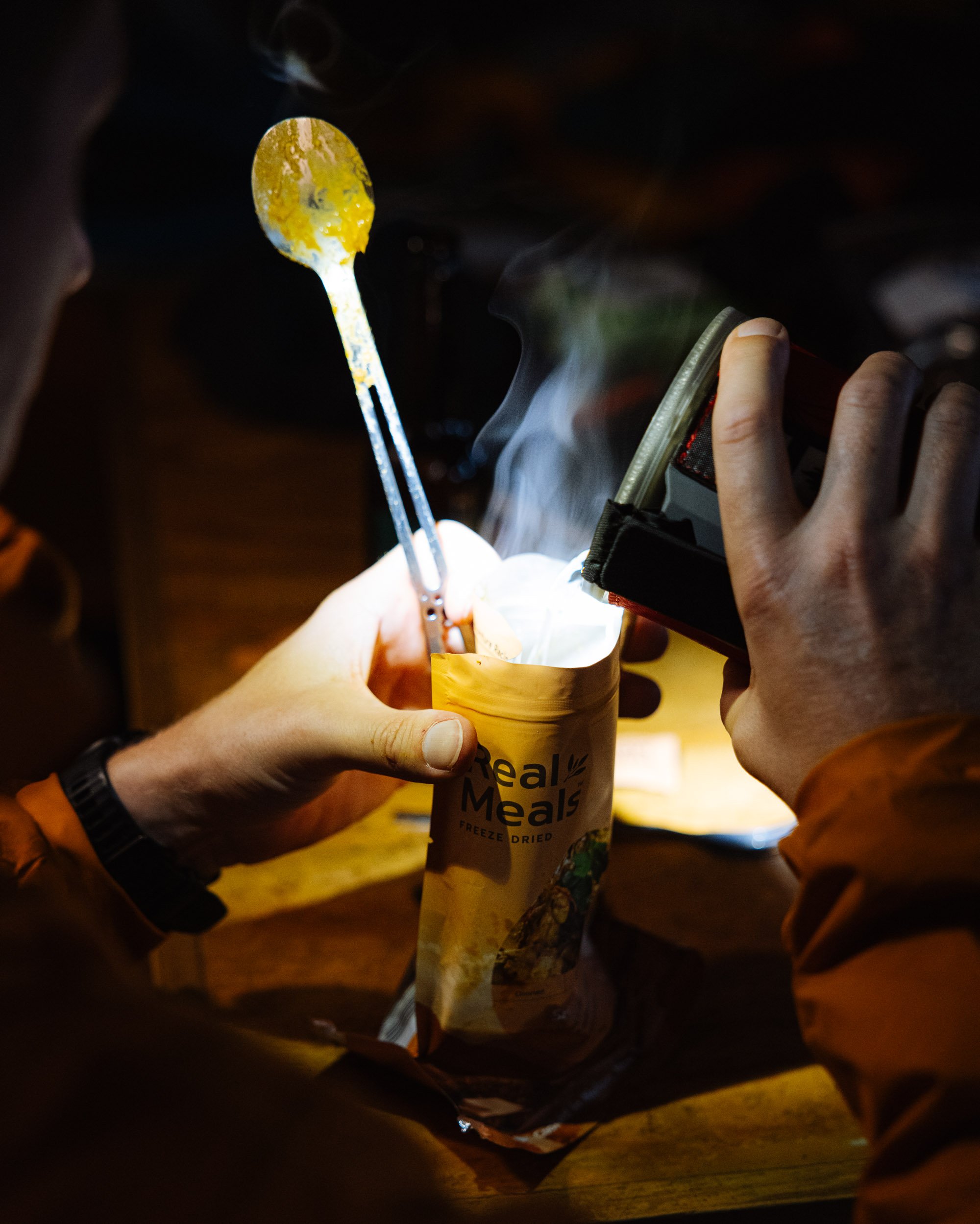 Person preparing freeze-dried meal with hot water, holding spoon and package on a table