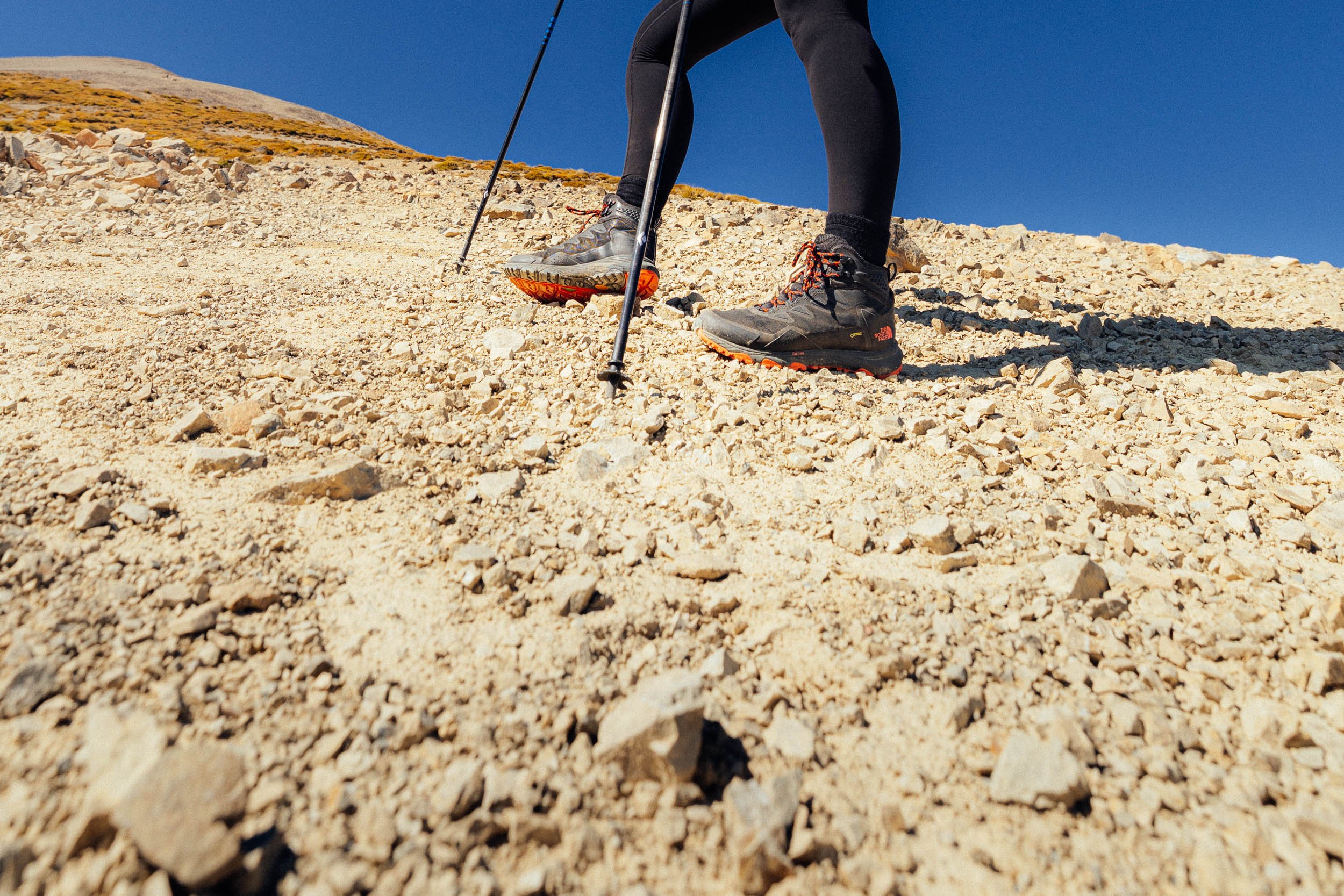 Person hiking on rocky terrain with trekking poles and hiking boots against a clear blue sky.