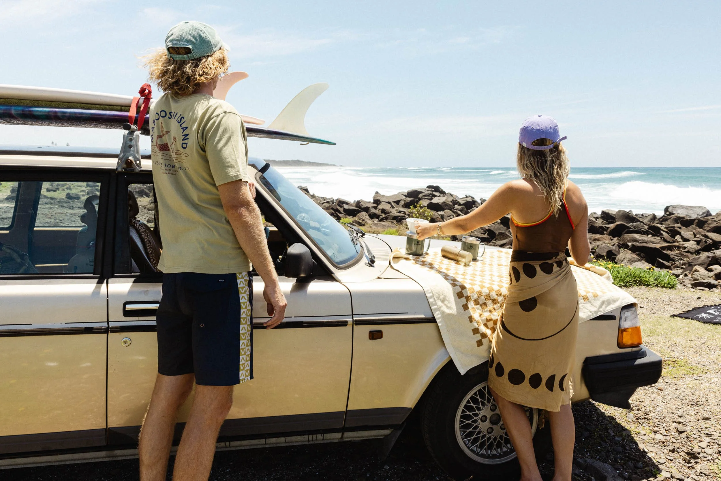 Two people by a vintage car with surfboards, on a beach with rocks and ocean waves in the background.