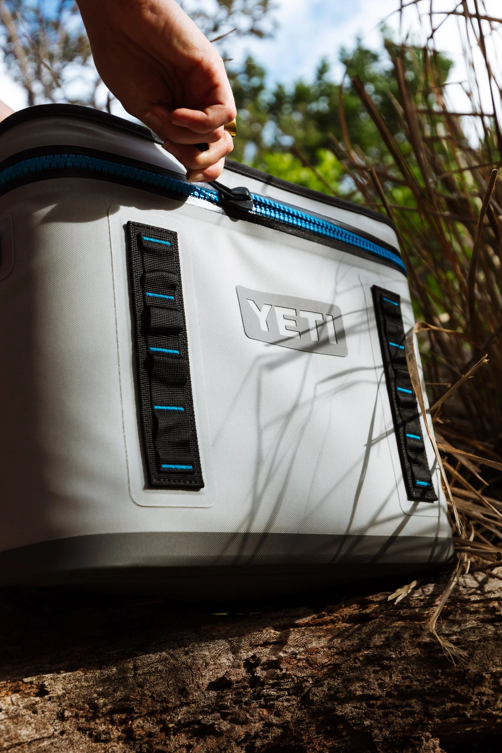 A hand reaching to open or close a YETI branded cooler or bag outdoors, with trees and a cloudy sky in the background.