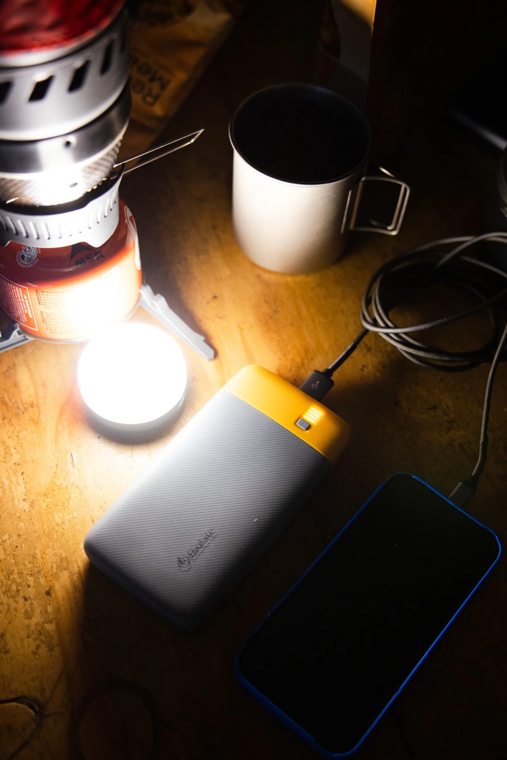 Camping setup with portable stove, mug, LED light, power bank, smartphone, and charging cable on a wooden surface.