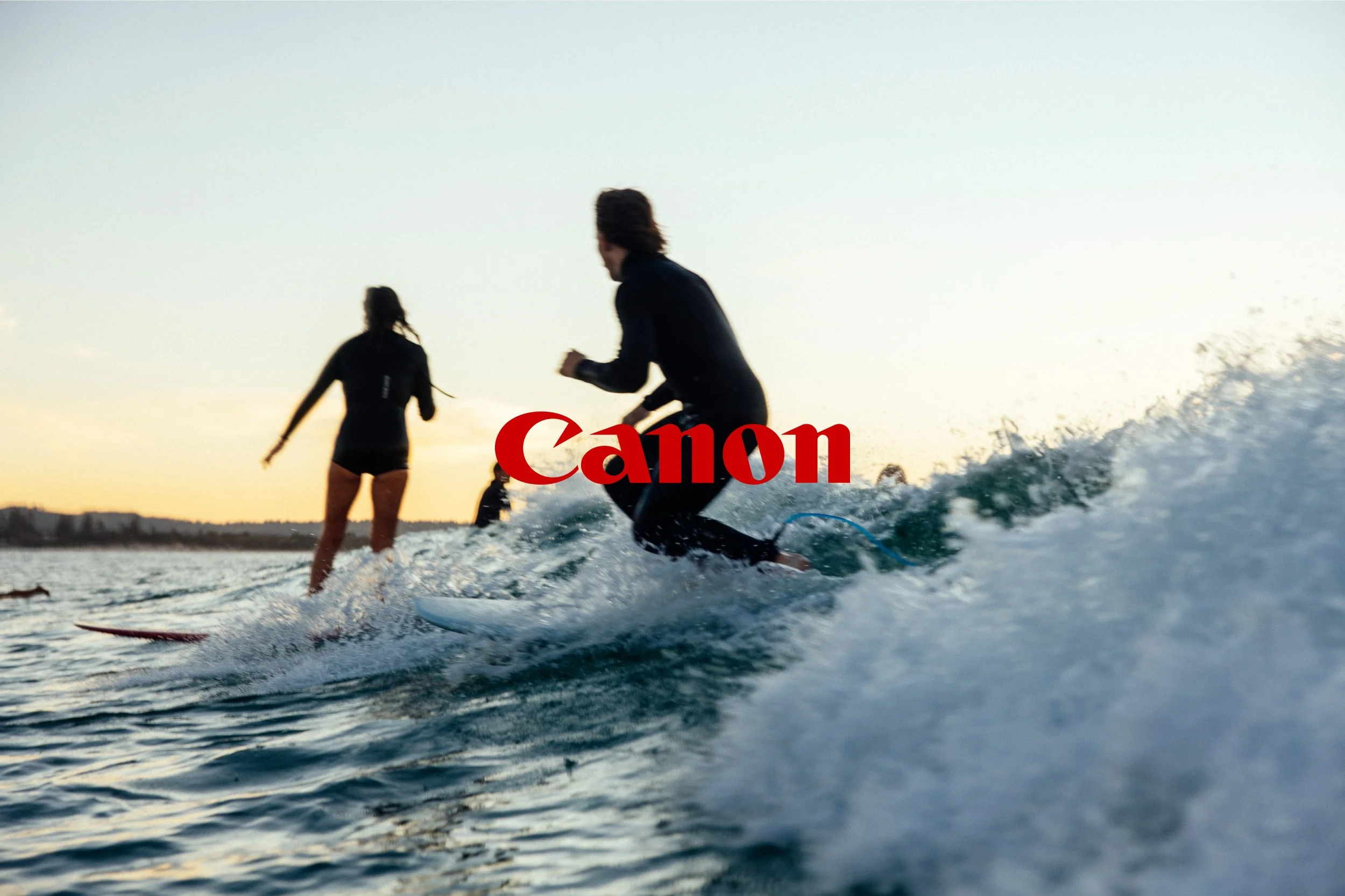 Surfers riding waves at sunset with Canon logo overlay.