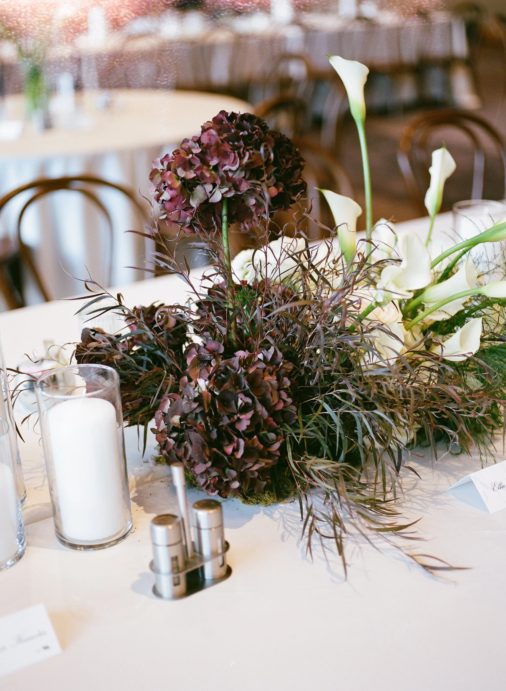 A floral centerpiece with dark purple and white flowers on a table with glass candle holders and silver votive candles, in a softly lit setting with wooden chairs in the background.
