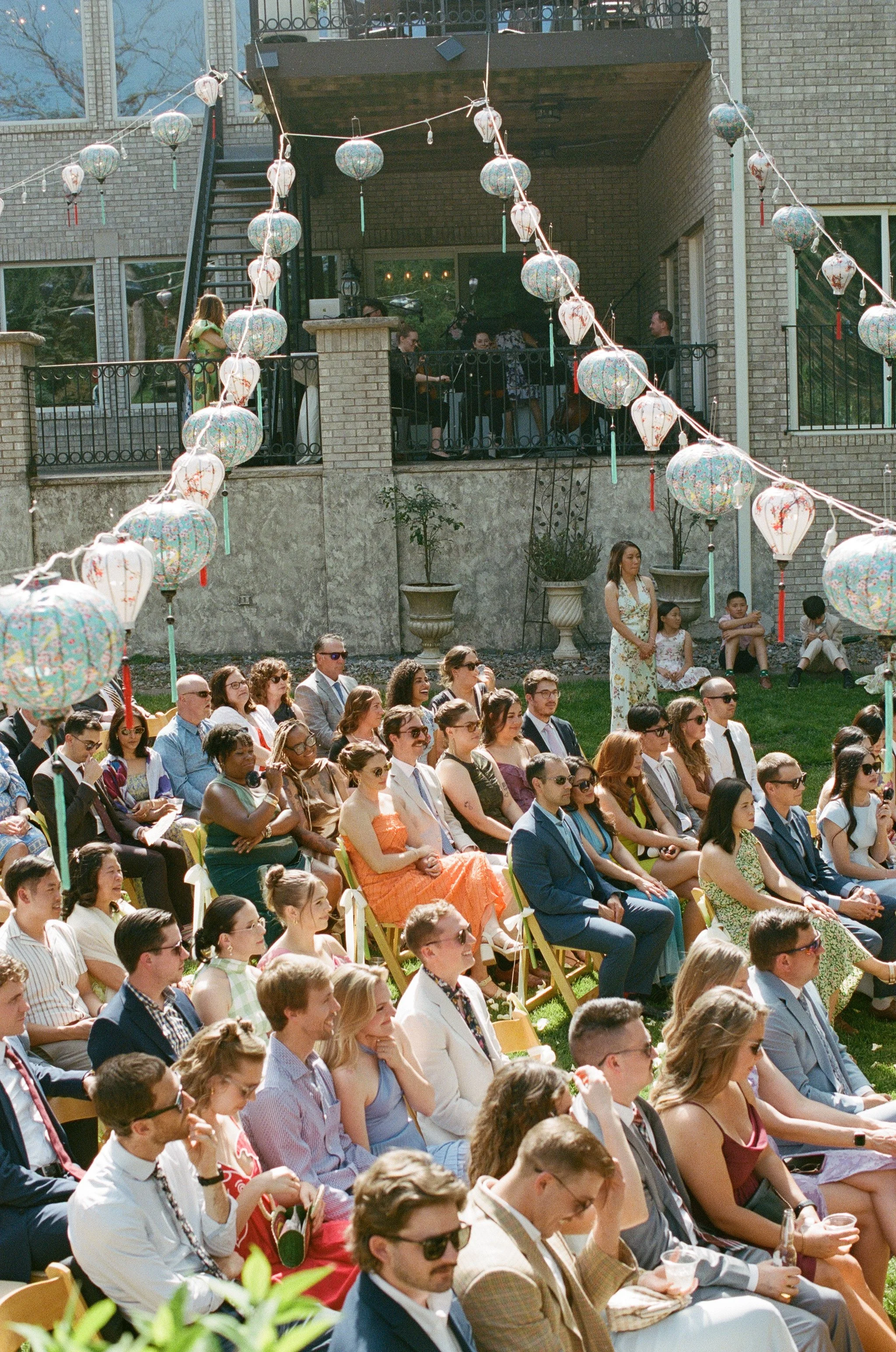 People attending an outdoor event with Chinese lanterns hanging overhead, seated and standing on the grass and on a patio.
