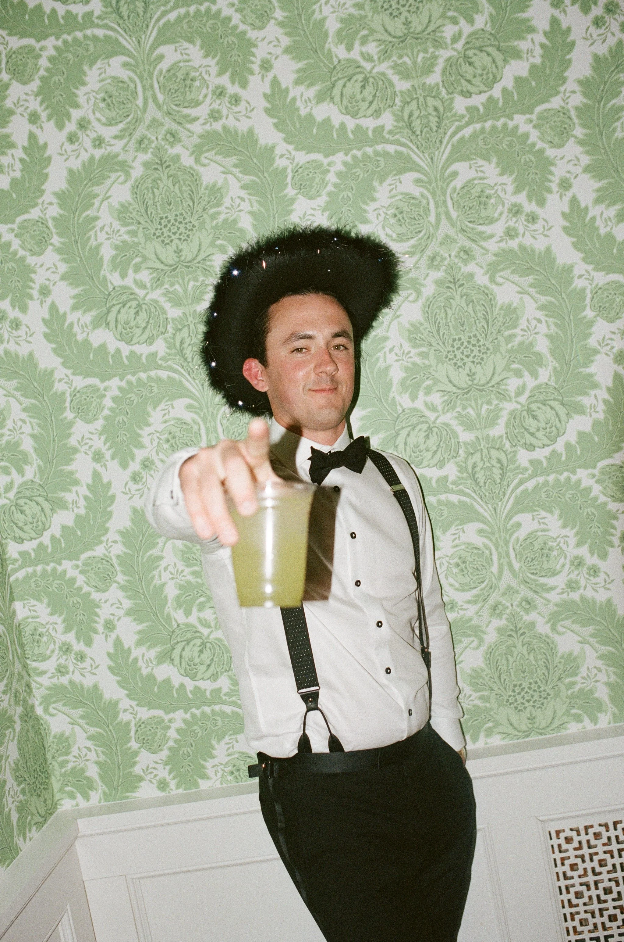 A man dressed in a tuxedo with suspenders and a bow tie is standing in front of a green floral wallpaper, pointing toward the camera with a confident expression, holding a glass of champagne.