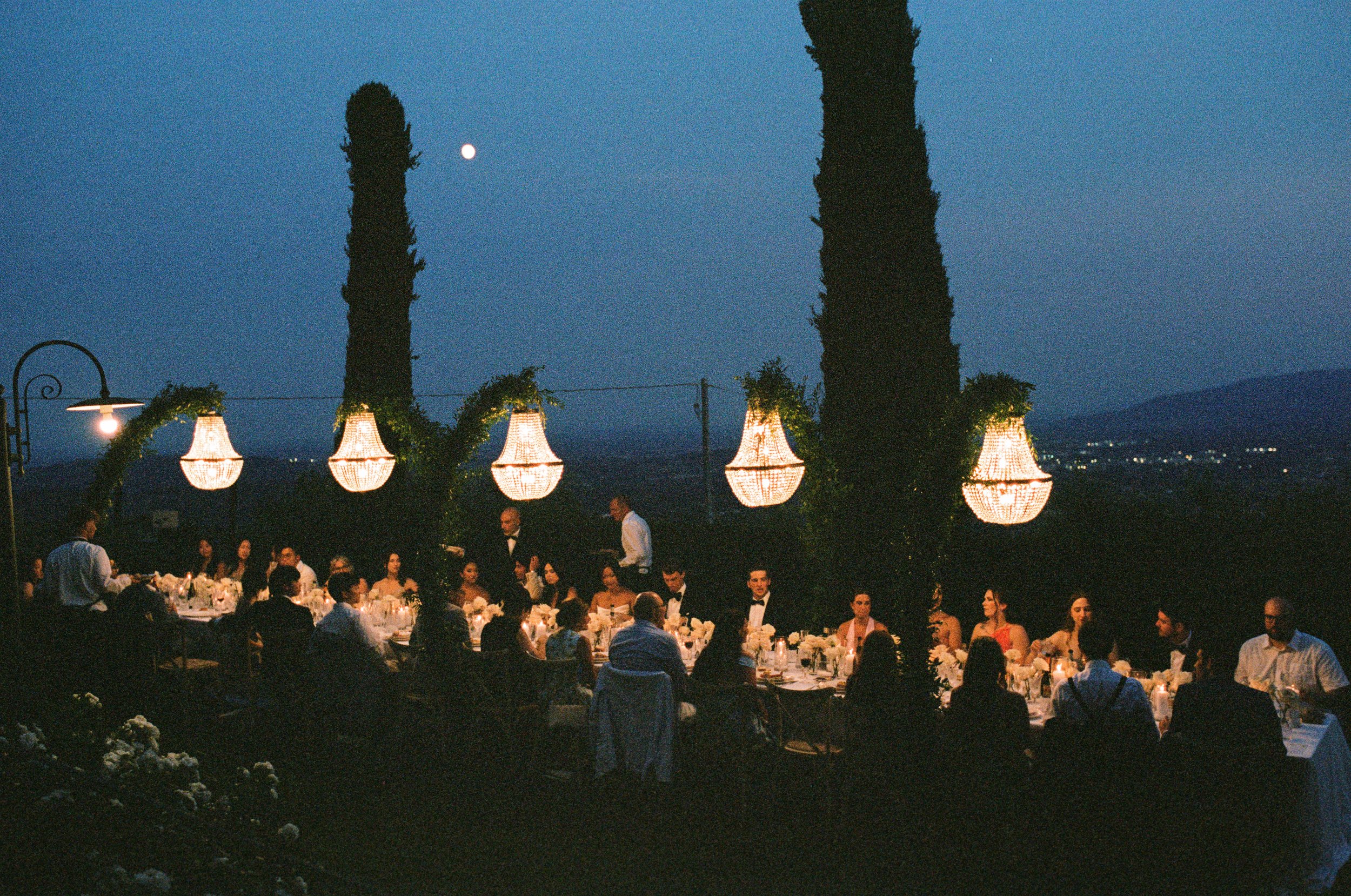 An outdoor evening event with guests dining at a long banquet table illuminated by hanging chandeliers, with tall trees and a cityscape in the background under a moonlit sky.