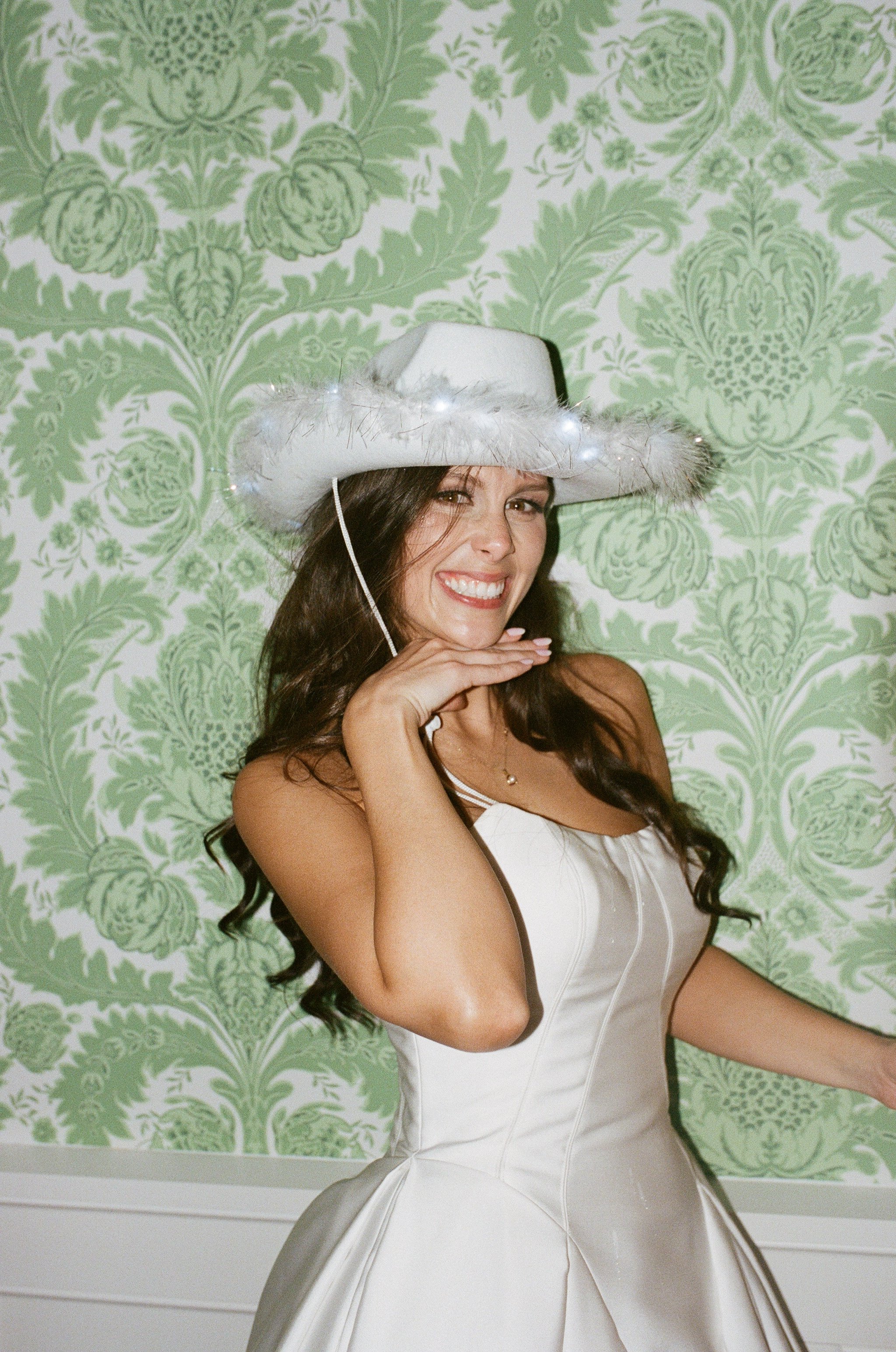 A woman with long dark hair wearing a white satin wedding dress and a cowboy hat decorated with feathers, smiling and posing in front of a green floral wallpaper background.