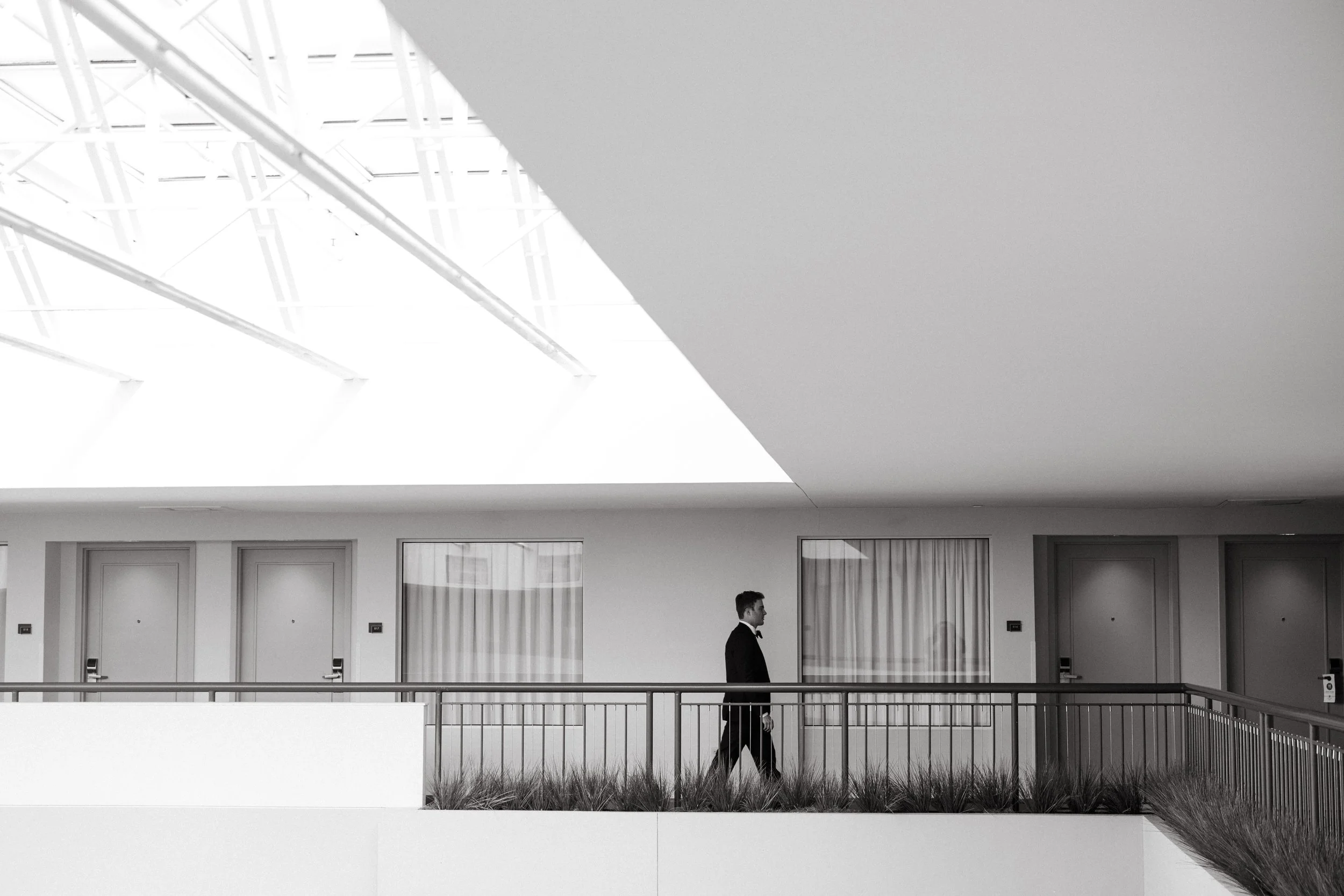 Black and white photo of a man walking indoors near railings, with a modern ceiling featuring skylights and structural beams.