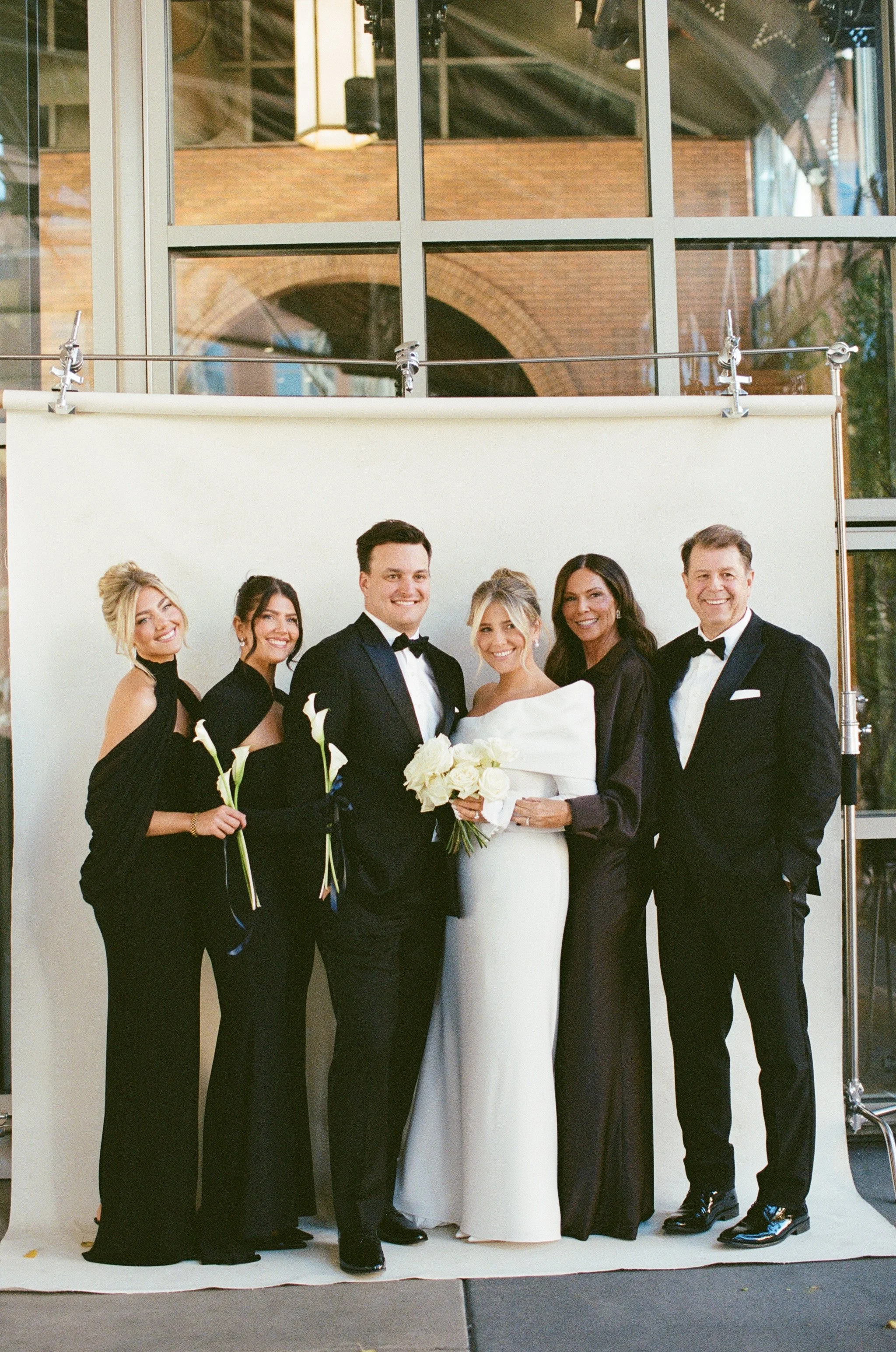 A wedding party poses in front of a white backdrop at a photography studio. The bride and groom are in the center, with the bride holding a bouquet and the groom wearing a tuxedo. Four bridesmaids and groomsmen stand around them, dressed in formal at