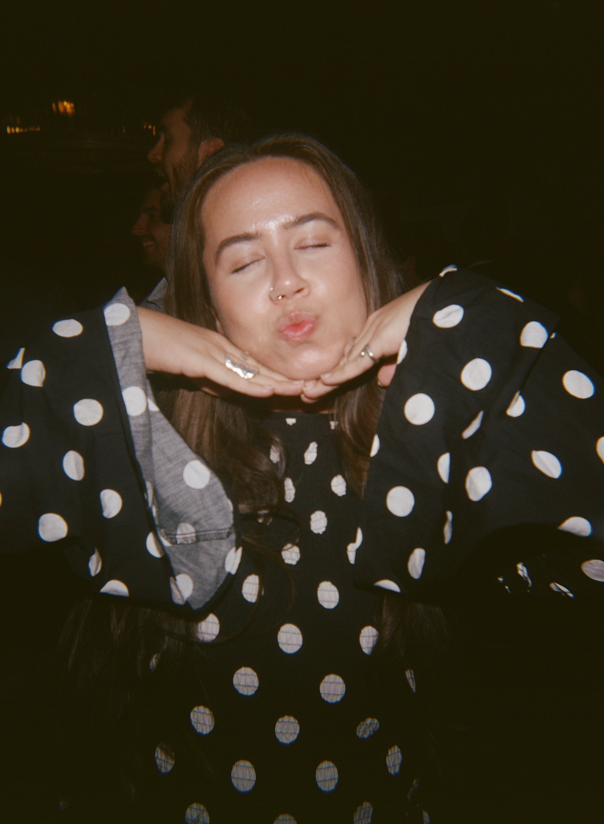 A woman with brown hair and a septum piercing pouting with her hands under her chin, wearing a black top with white polka dots. Behind her are two men, one with dark hair and beard, the other with short hair, both looking away into the distance against a dark background.