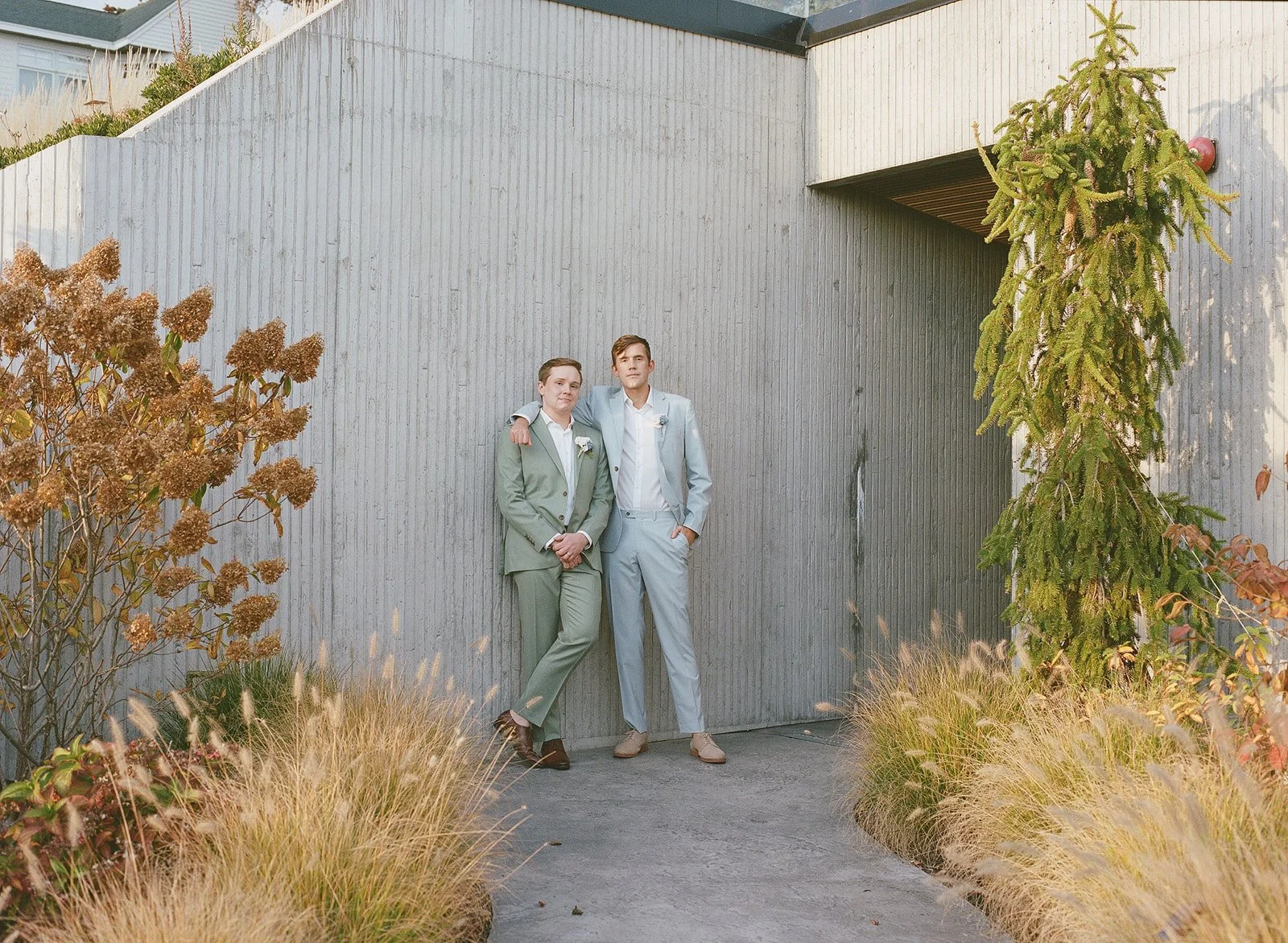 Two young men in suits standing outside near a modern wooden building, with plants and trees around, one has his arm around the other's shoulder.