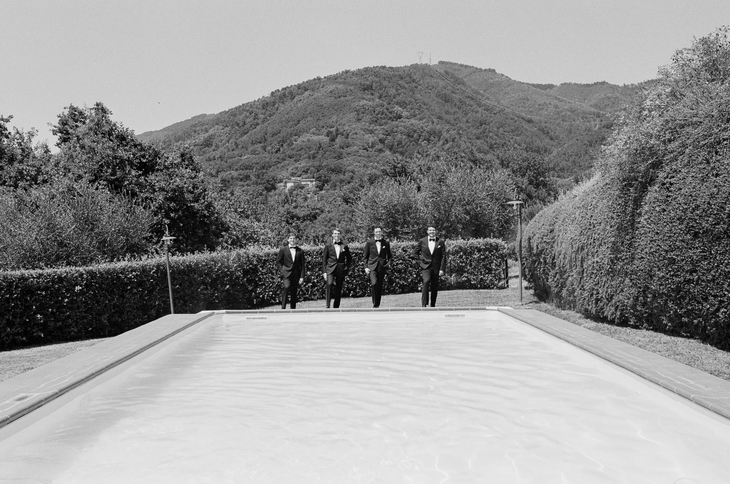 Four men dressed in tuxedos walking outdoors in front of a scenic hill with trees and a mountain, with a pool or water feature in the foreground.