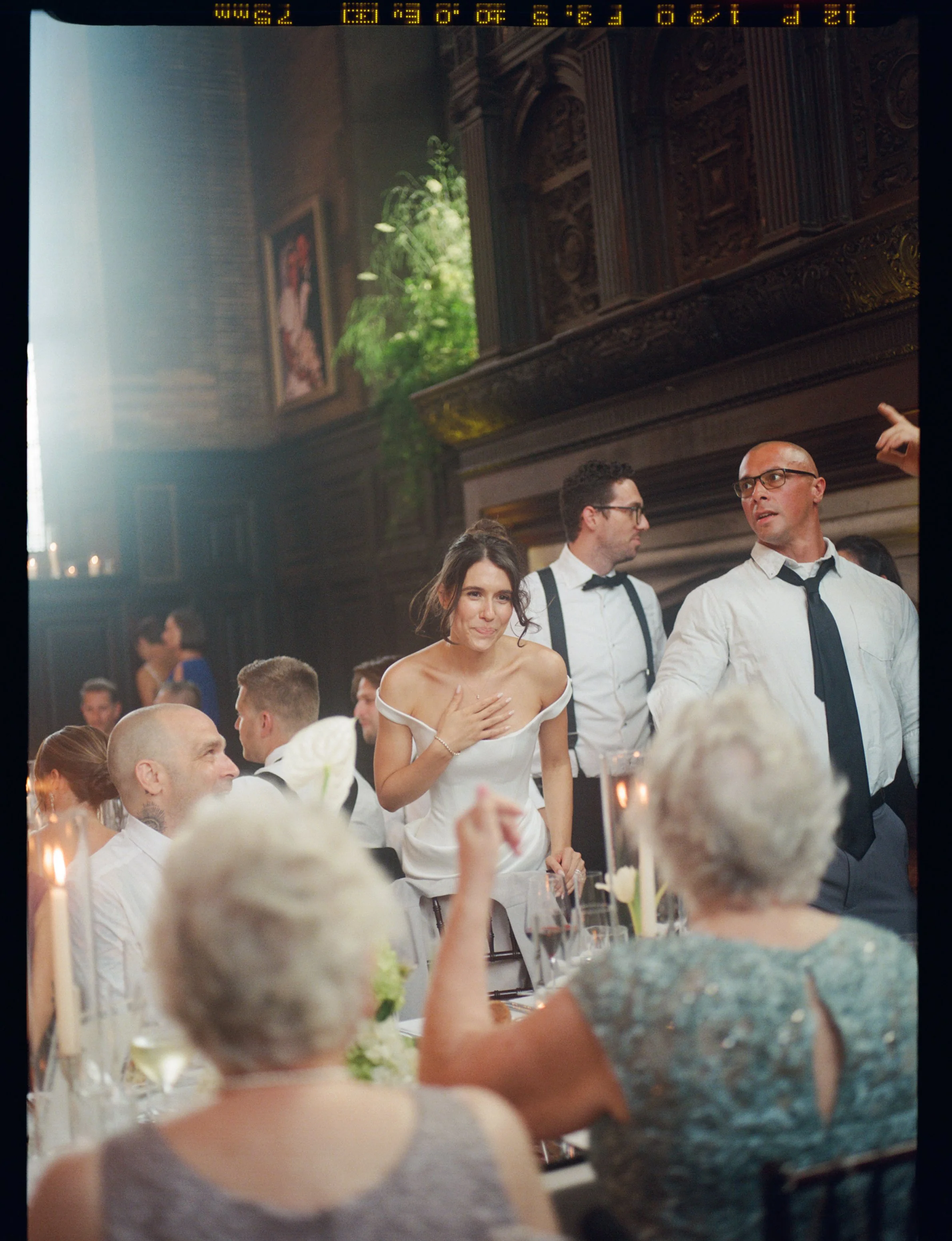 A woman in a white dress touching her chest at an elegant dinner event with many guests, ornate woodwork on the walls, and framed artwork, in a luxurious historic setting.