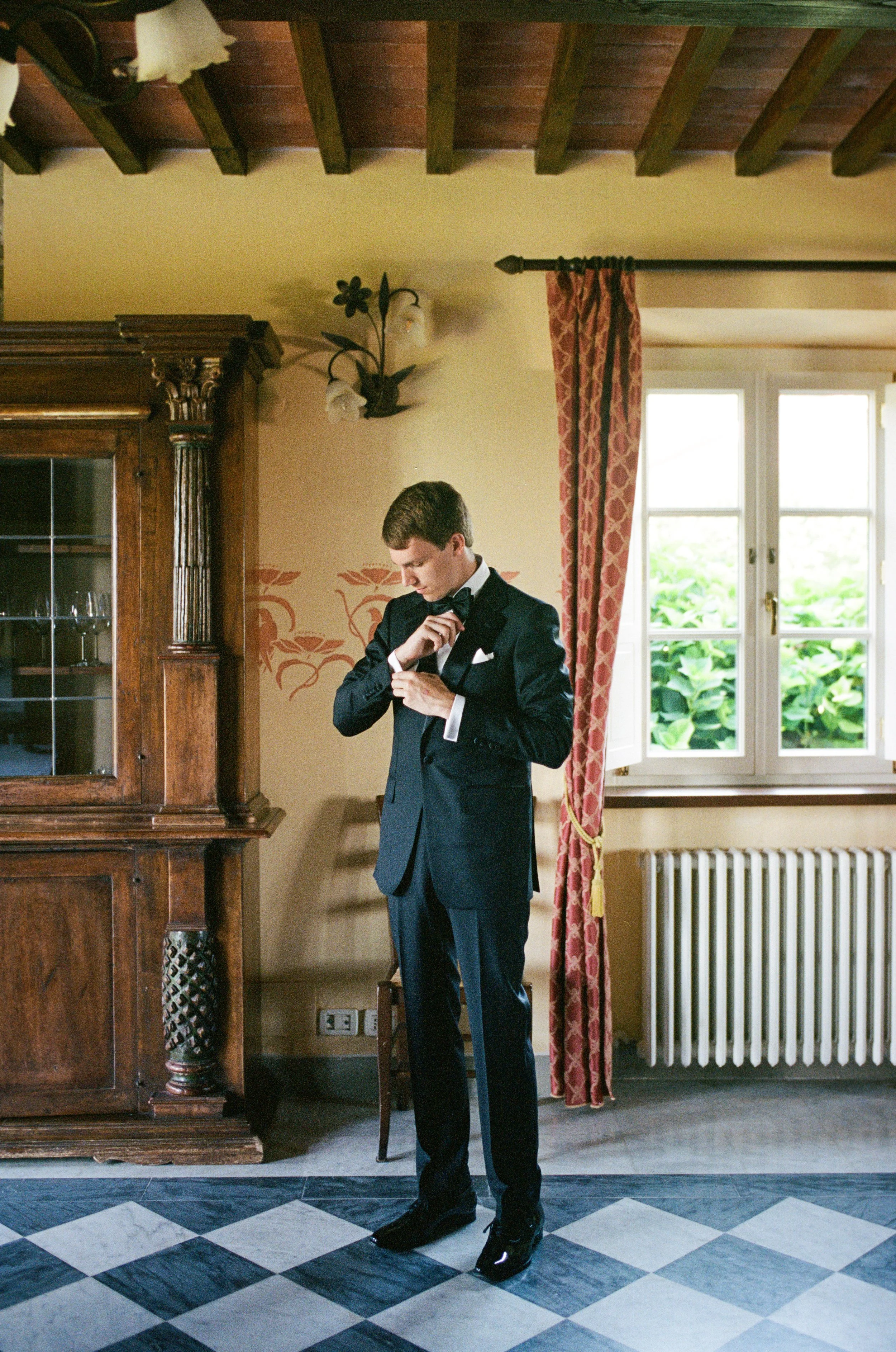 A man in a formal black tuxedo standing in a vintage styled room adjusting his bow tie. The room has a window with red patterned curtains, a wooden cabinet with glass doors, a wall light fixture, and a tiled floor with a black and white checkered pat
