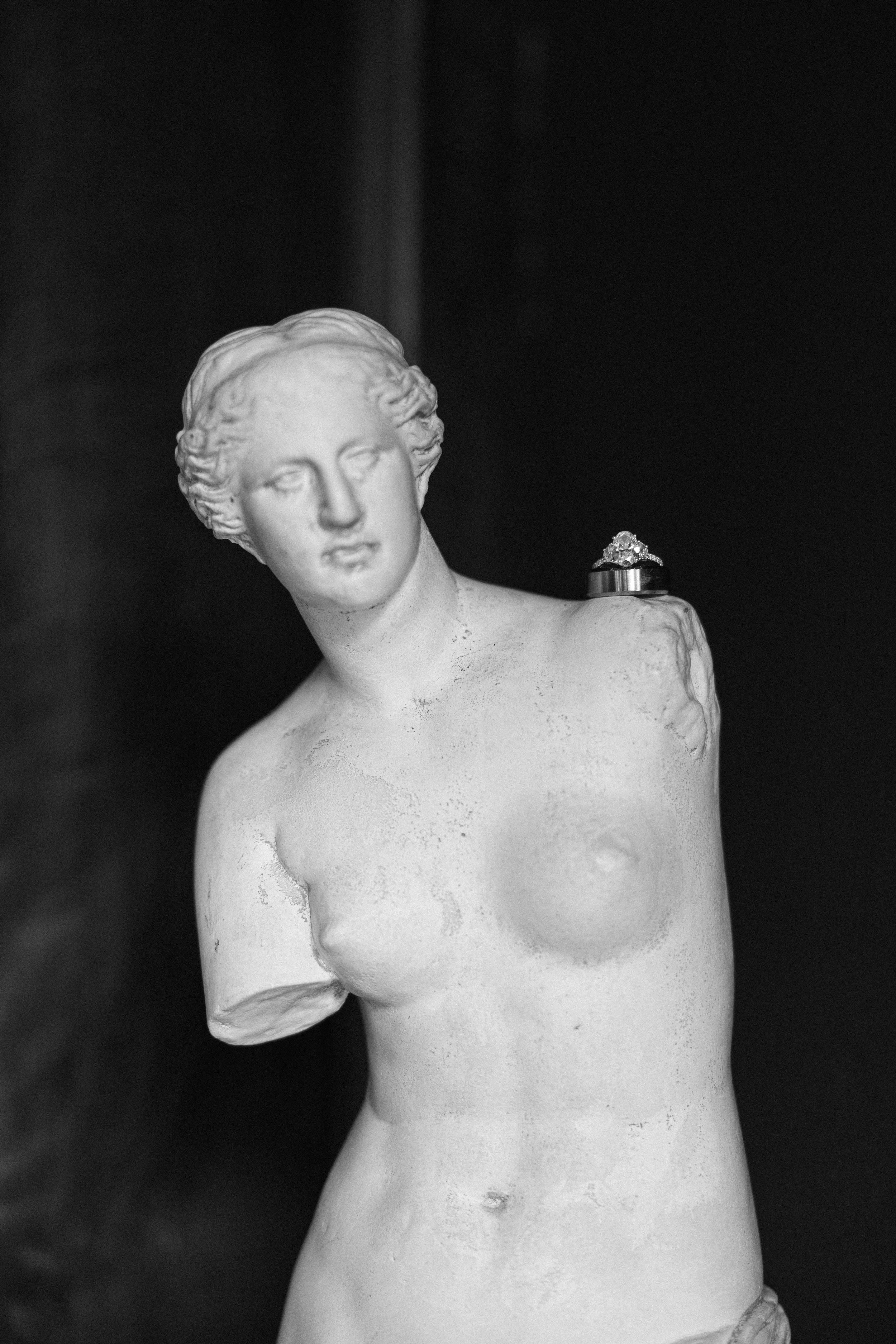 Black and white photograph of a classical style female sculpture's bust, featuring detailed hair and facial features, with a diamond ring resting on her shoulder.