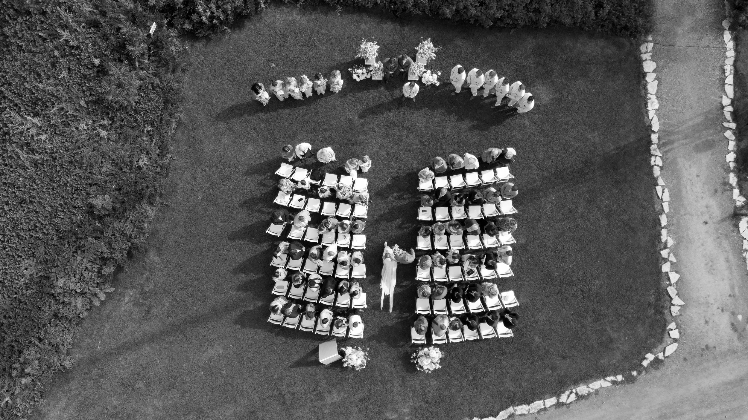 Black and white aerial view of a wedding ceremony outdoors with guests seated in rows, a bride and groom in the center, and flower arrangements.