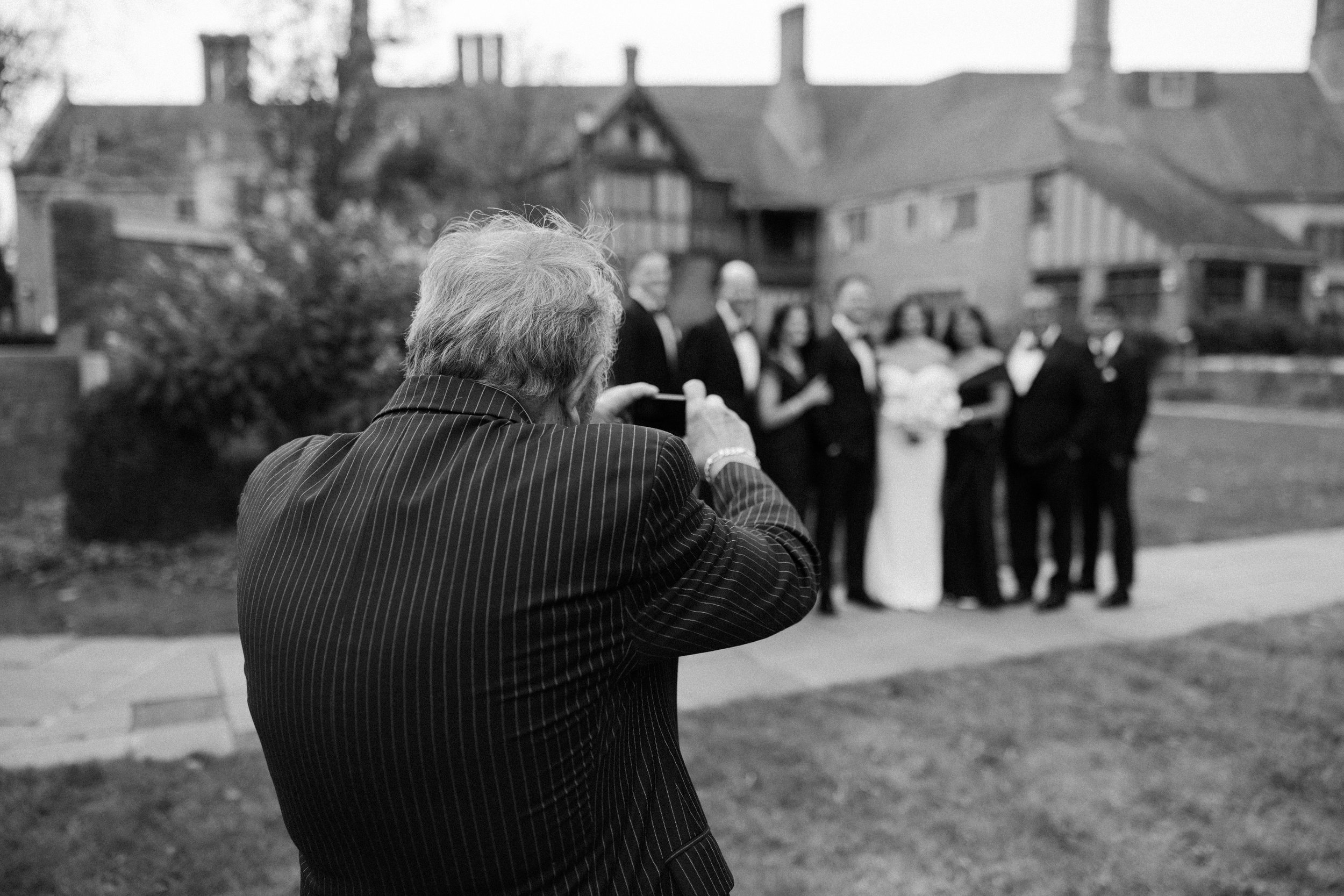 A man with gray hair wearing a pinstripe suit filming a wedding party of five people outside on a lawn, with houses in the background.