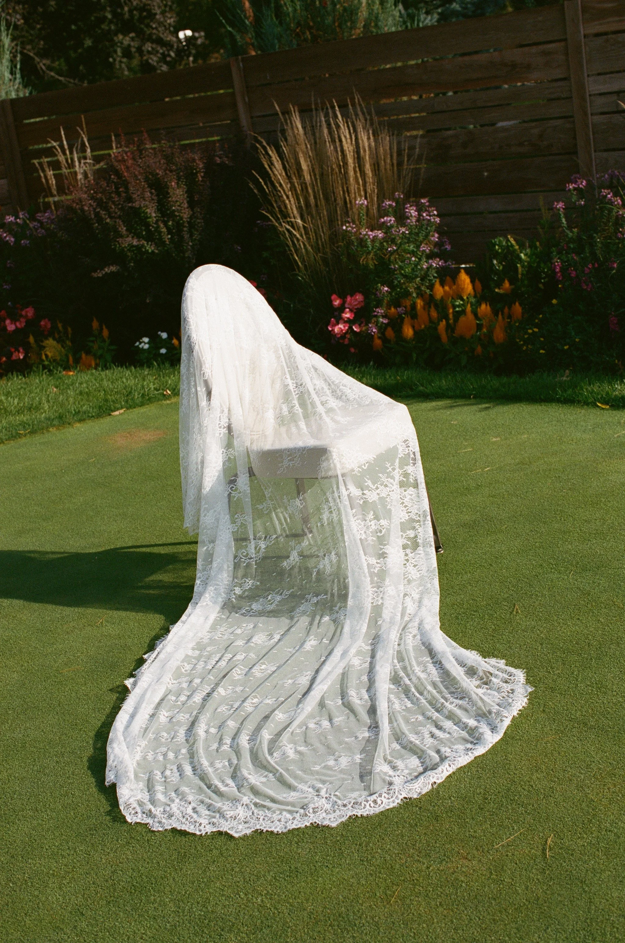 A white lace wedding veil draped over a chair on a grassy lawn in front of a flower garden and wooden fence.