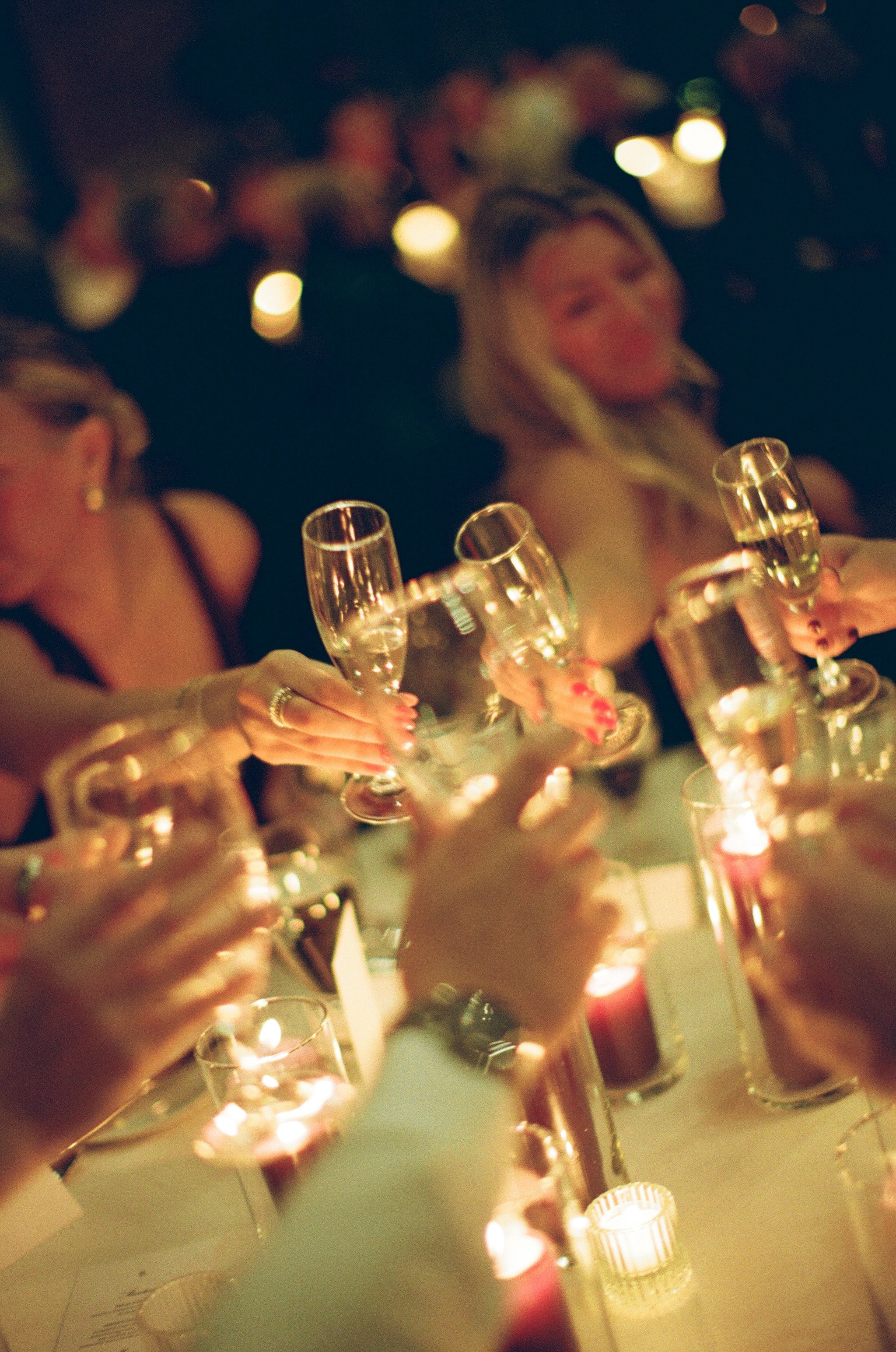 Women celebrating with champagne glasses in a dimly lit setting with candles and blurred background lights.