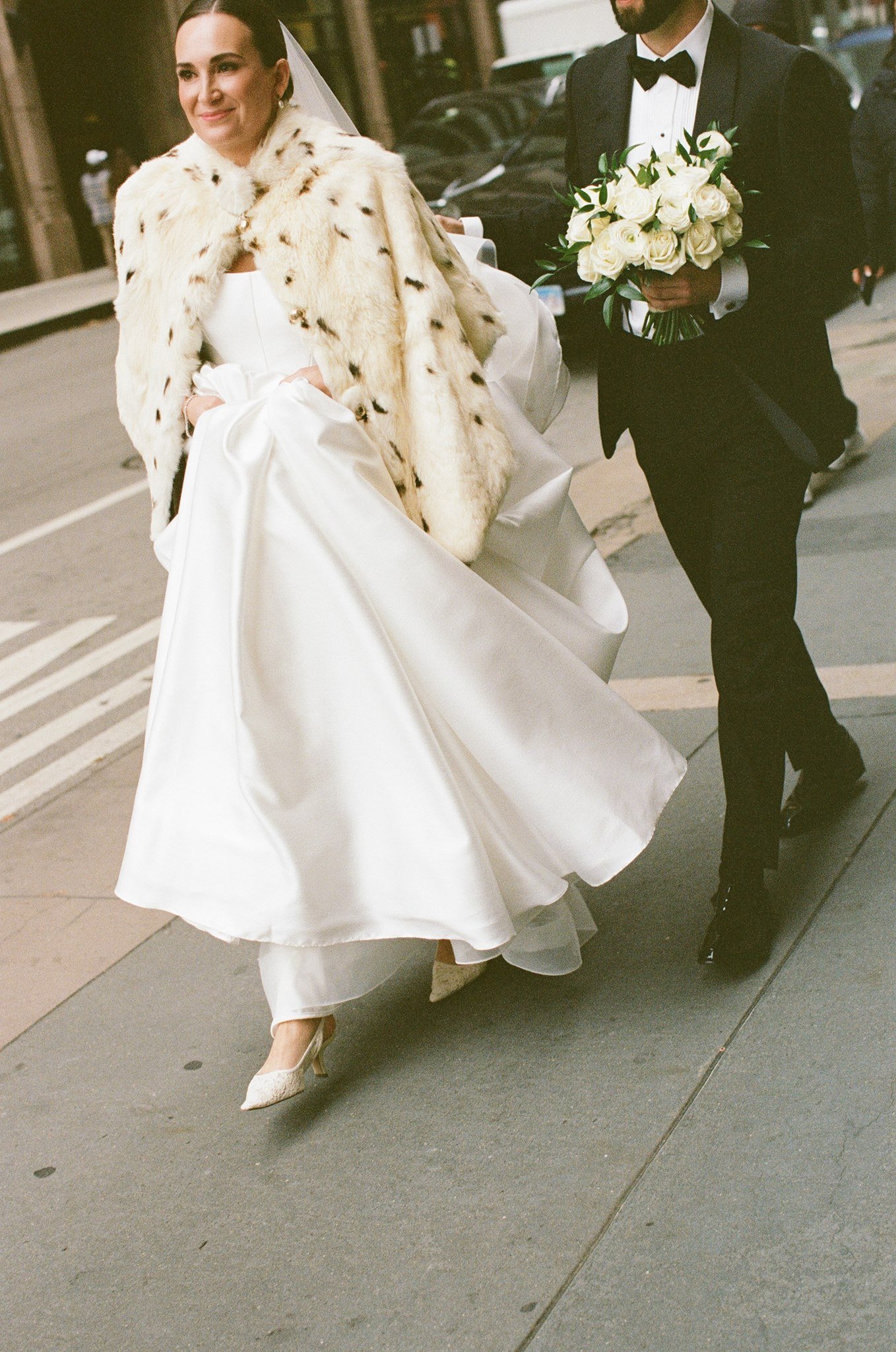 A bride in a white wedding dress with a fur cape walking on a city sidewalk, holding up her dress, being followed by a man in a tuxedo carrying a bouquet of white roses.