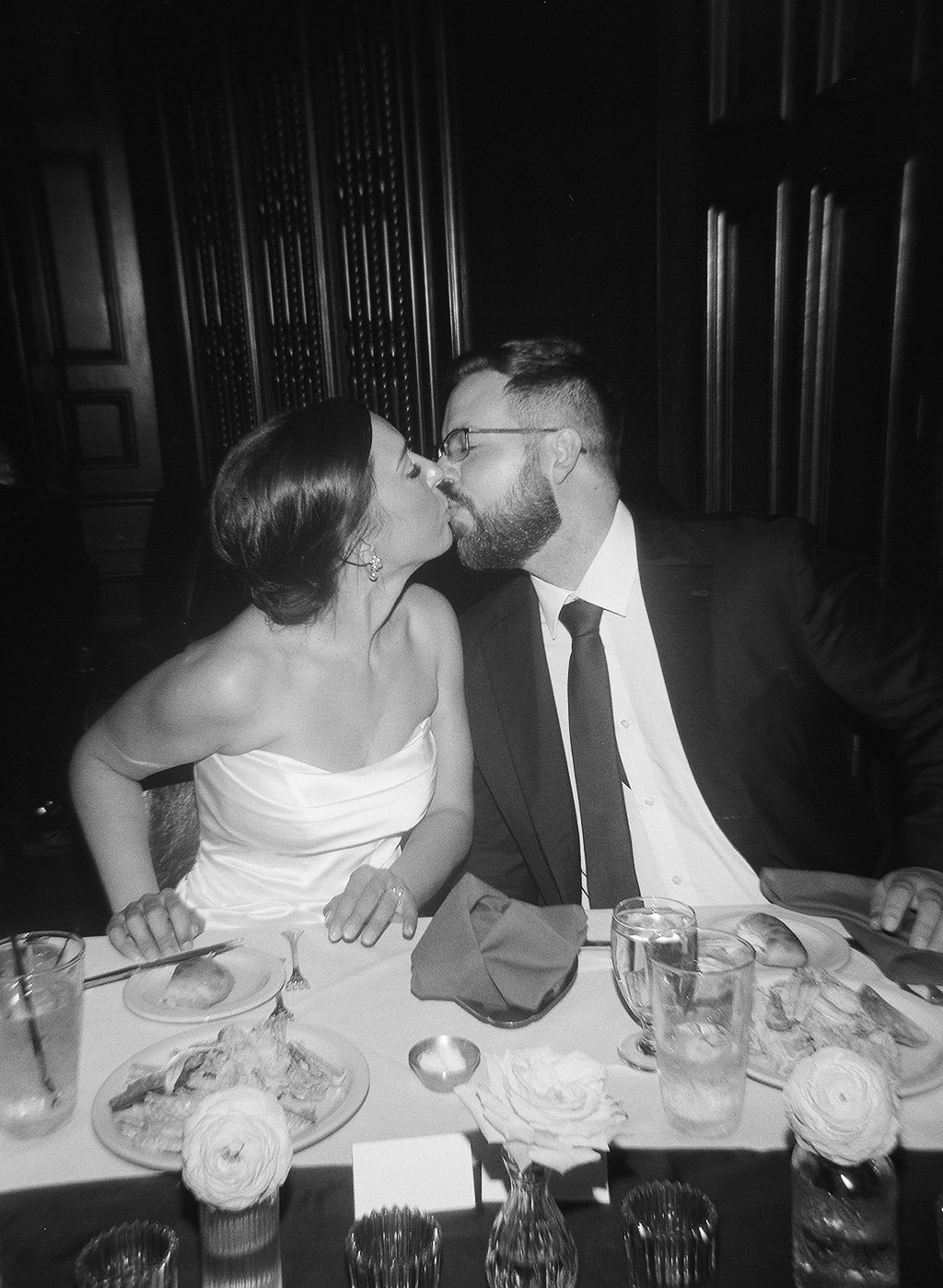 A black and white photo of a woman and a man kissing at a formal event. The woman is wearing a strapless dress, and the man is in a suit with glasses. They are seated at a table with food, drinks, and flowers.