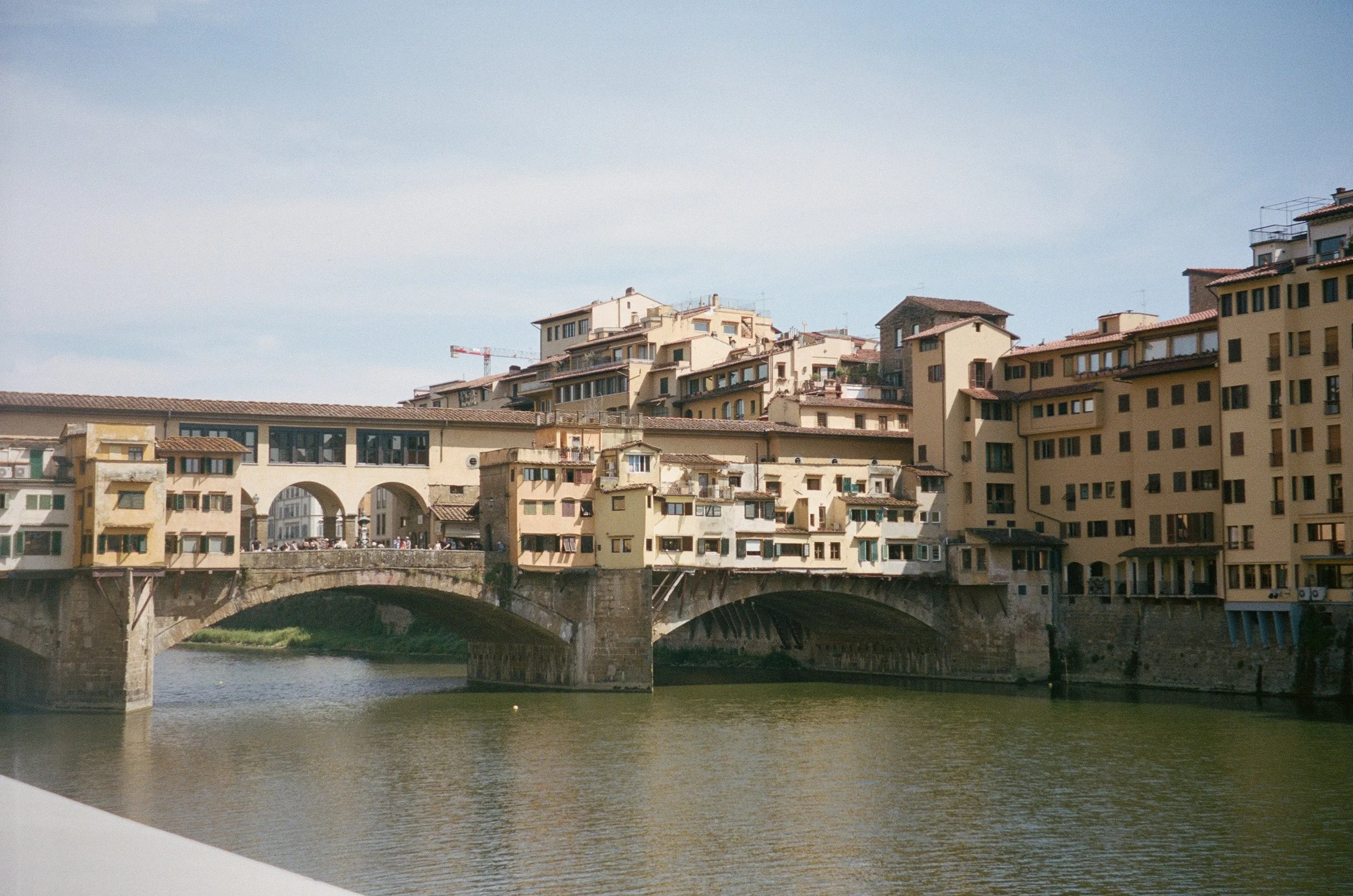 View of a historic bridge with multiple arches passing over a river, with a cluster of pastel-colored buildings with terracotta roofs on a hill in the background under a clear blue sky.